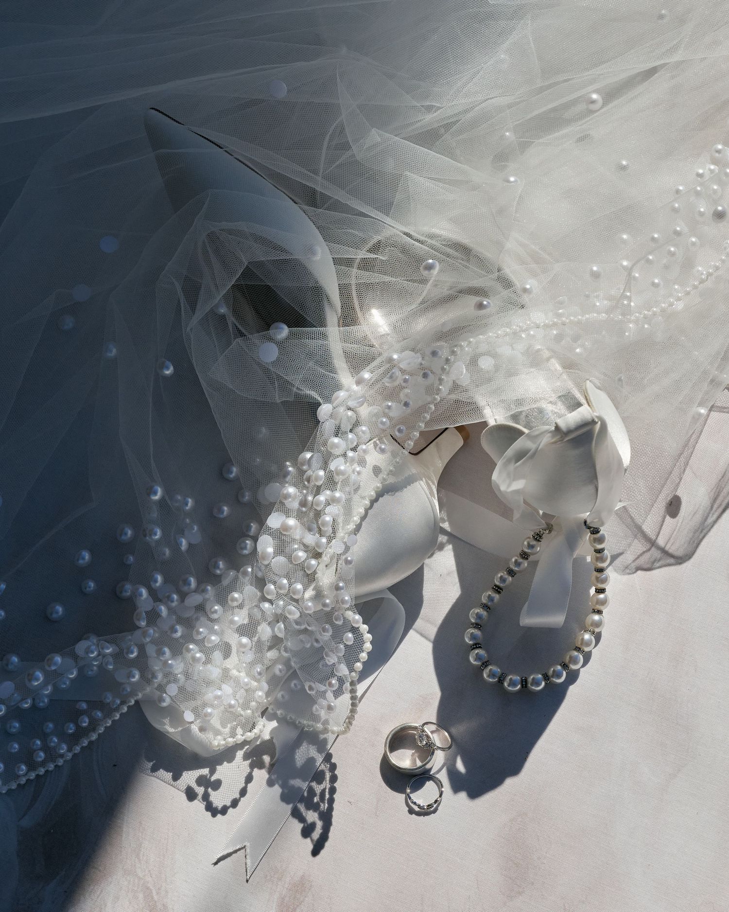 Wedding accessories including a delicate veil, pearl necklace, and diamond rings arranged on a light surface.