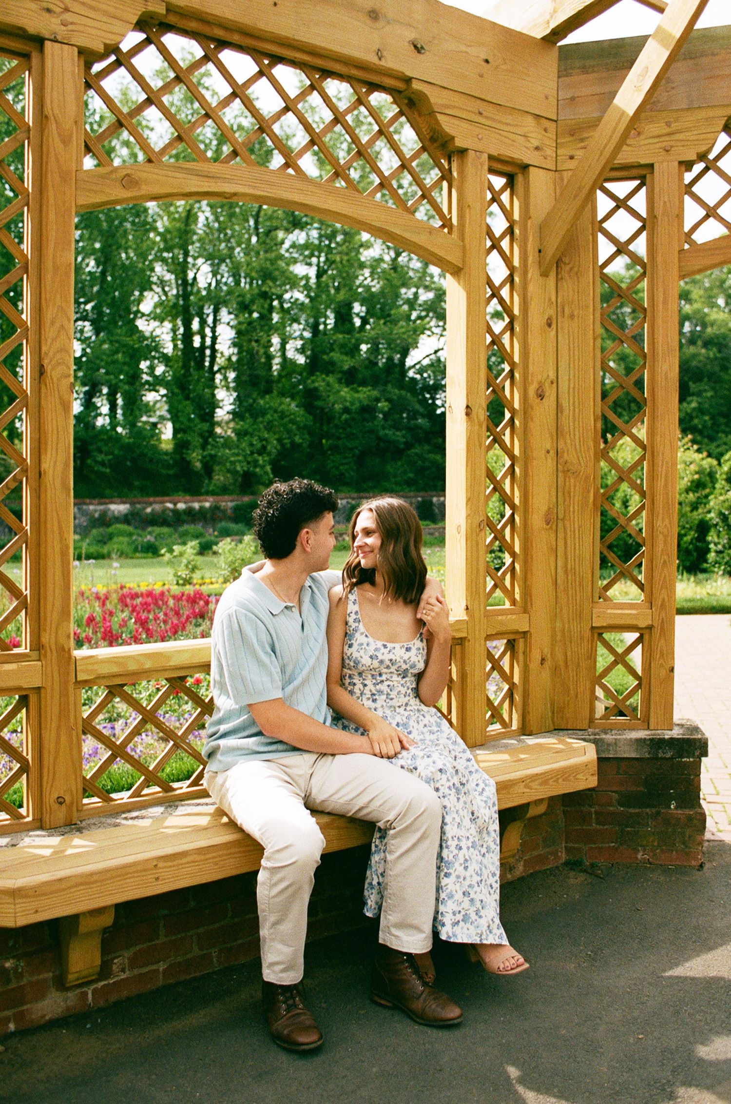 Couple sitting together on a wooden bench inside a garden pergola, smiling at each other surrounded by lush greenery.