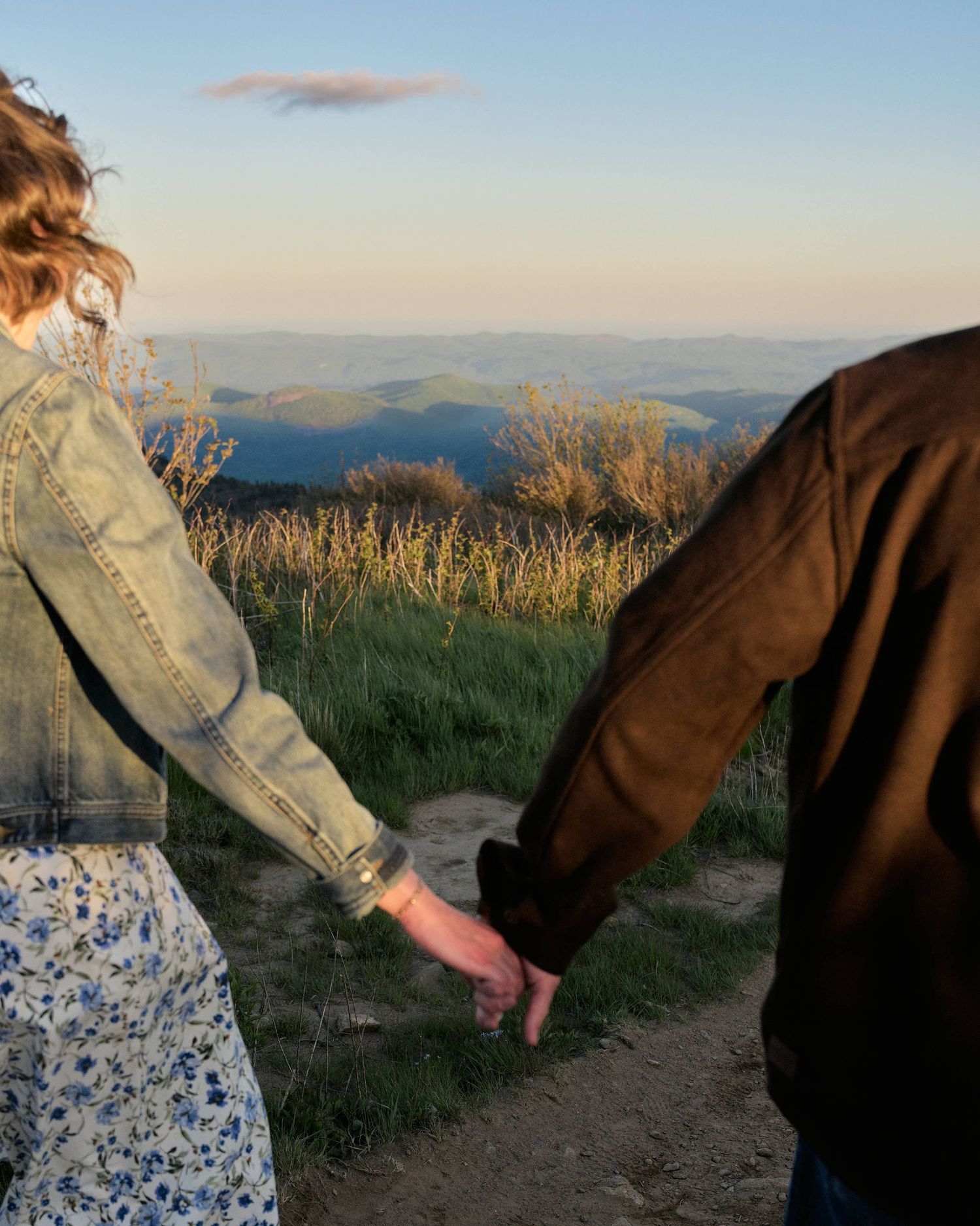 Couple holding hands on a hilltop at golden hour, overlooking rolling green mountains under a clear sky.