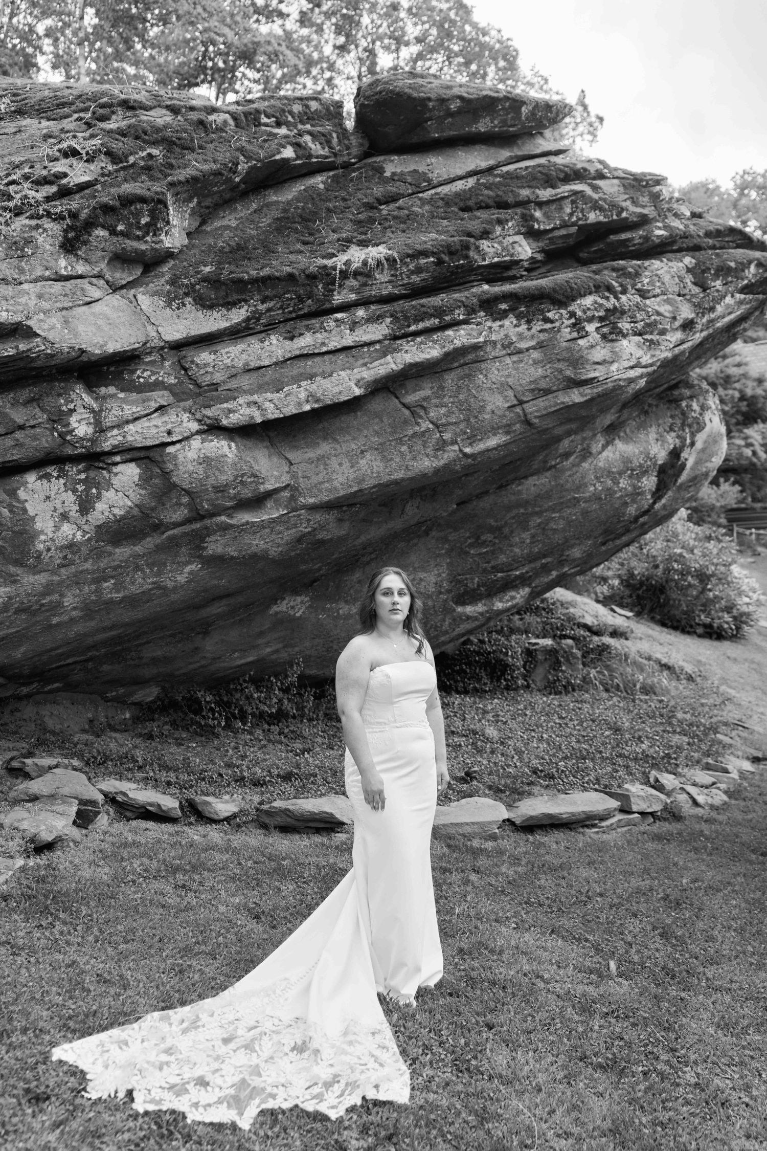 Bride in white gown with lace train stands before large rock formation in black and white outdoor bridal portrait.