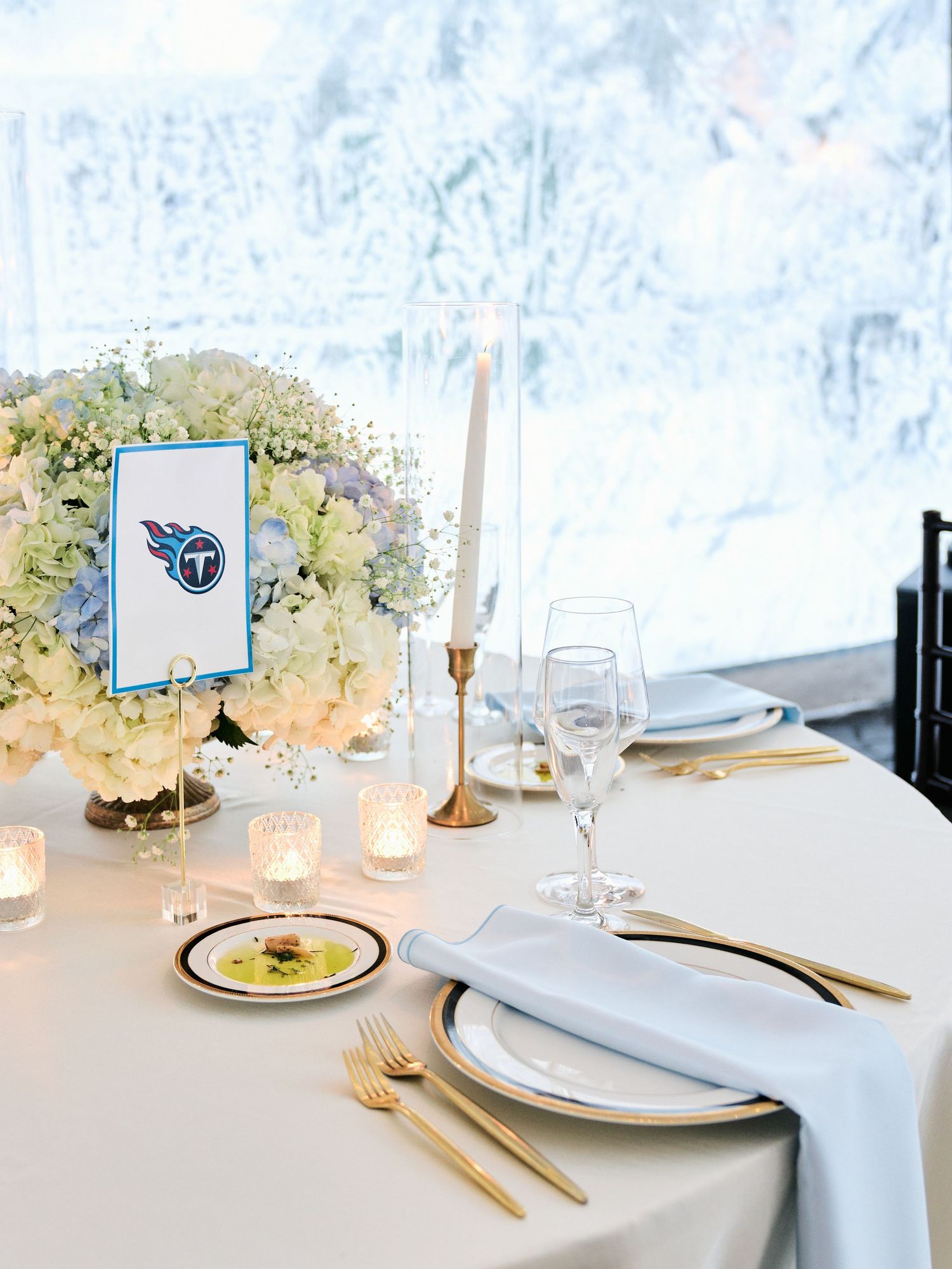 Elegant Tennessee Titans-themed wedding table setting with gold cutlery, white florals, and candles against a snowy backdrop.