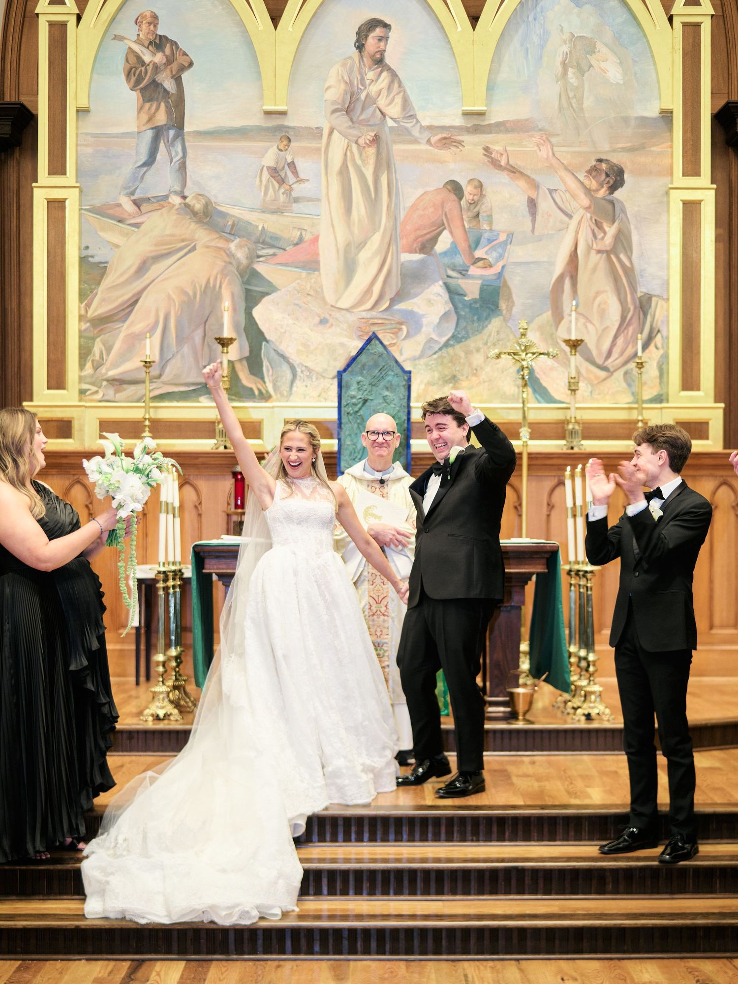Joyful bride and groom celebrate at church altar with raised arms, officiant and wedding party, beneath a religious mural.