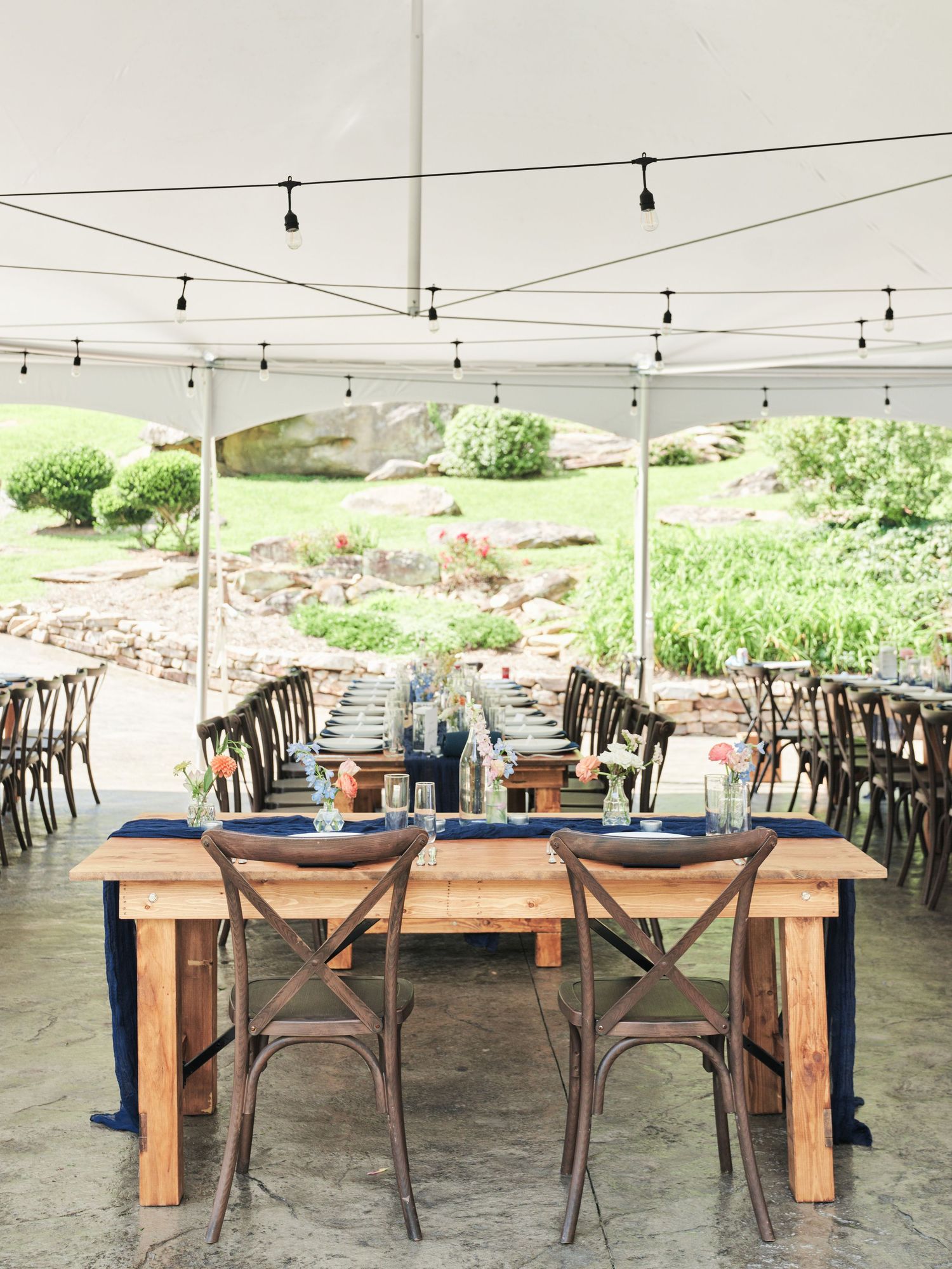 Elegant outdoor wedding reception under a white tent with rustic wood farm tables, cross-back chairs, and navy blue table runners.