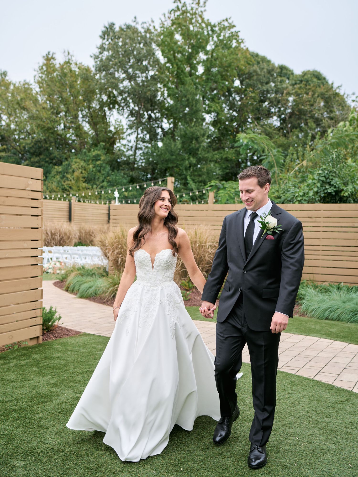 A bride in a white ball gown and groom in a dark suit hold hands walking through a garden wedding venue.