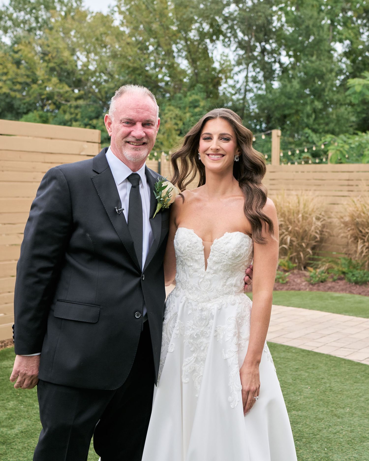 Bride in white lace gown poses with older man in black suit and tie outdoors at wedding venue.