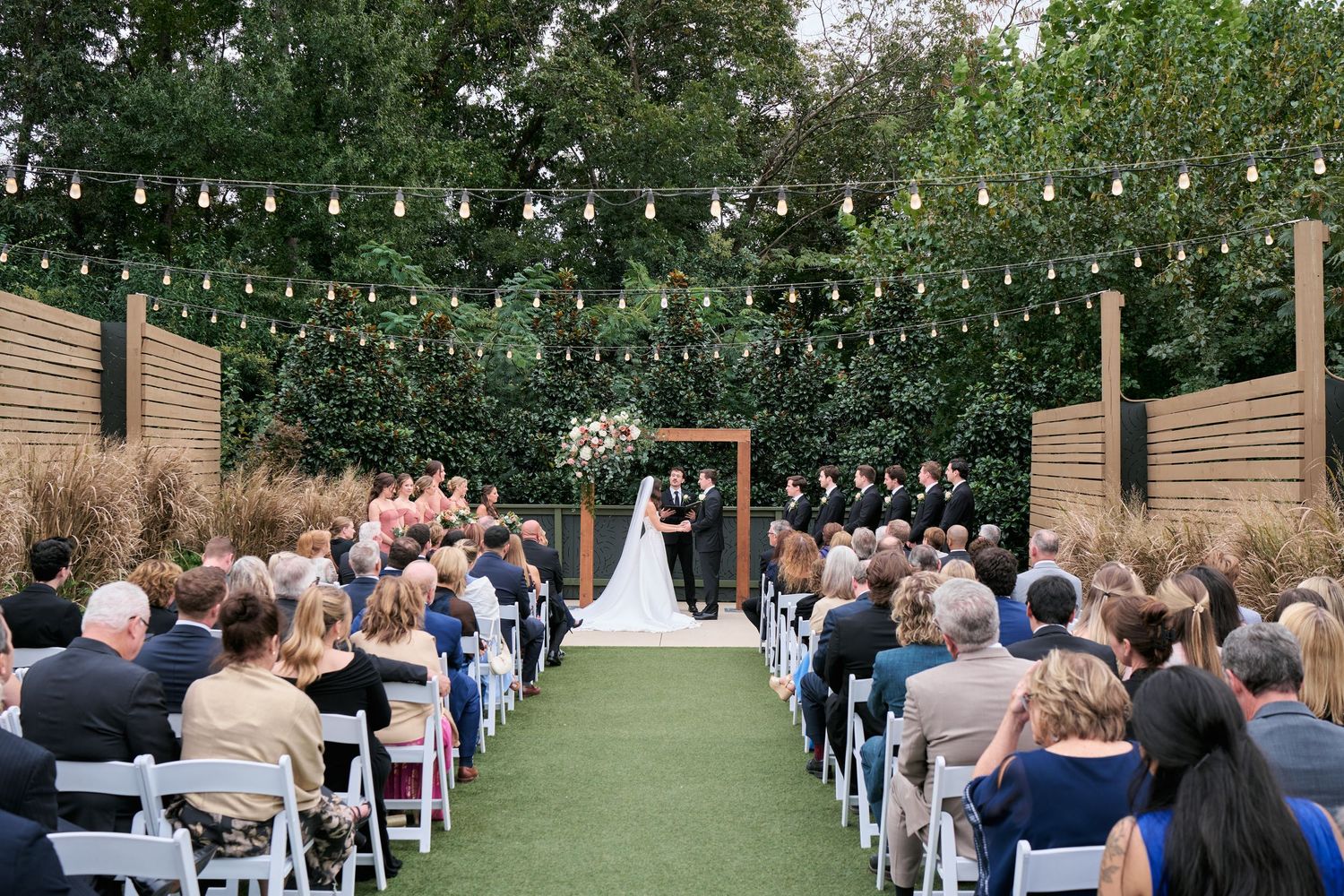 Outdoor wedding ceremony with string lights, white chairs, and guests watching couple at floral arch amid lush greenery.