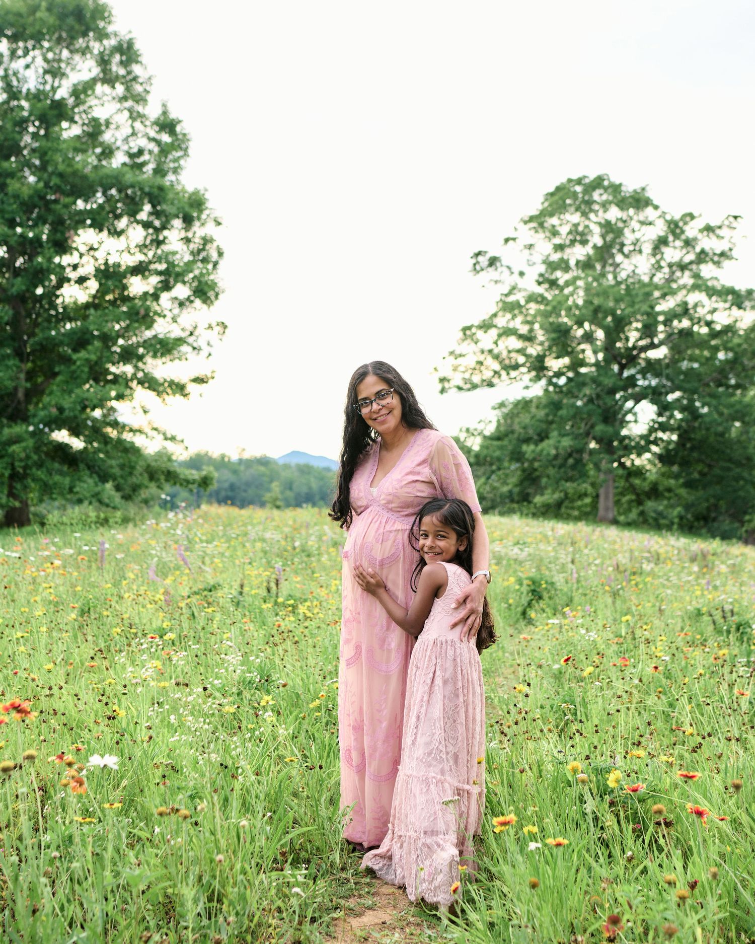 Pregnant woman in pink maxi dress hugging young daughter in floral field with wildflowers and green trees.
