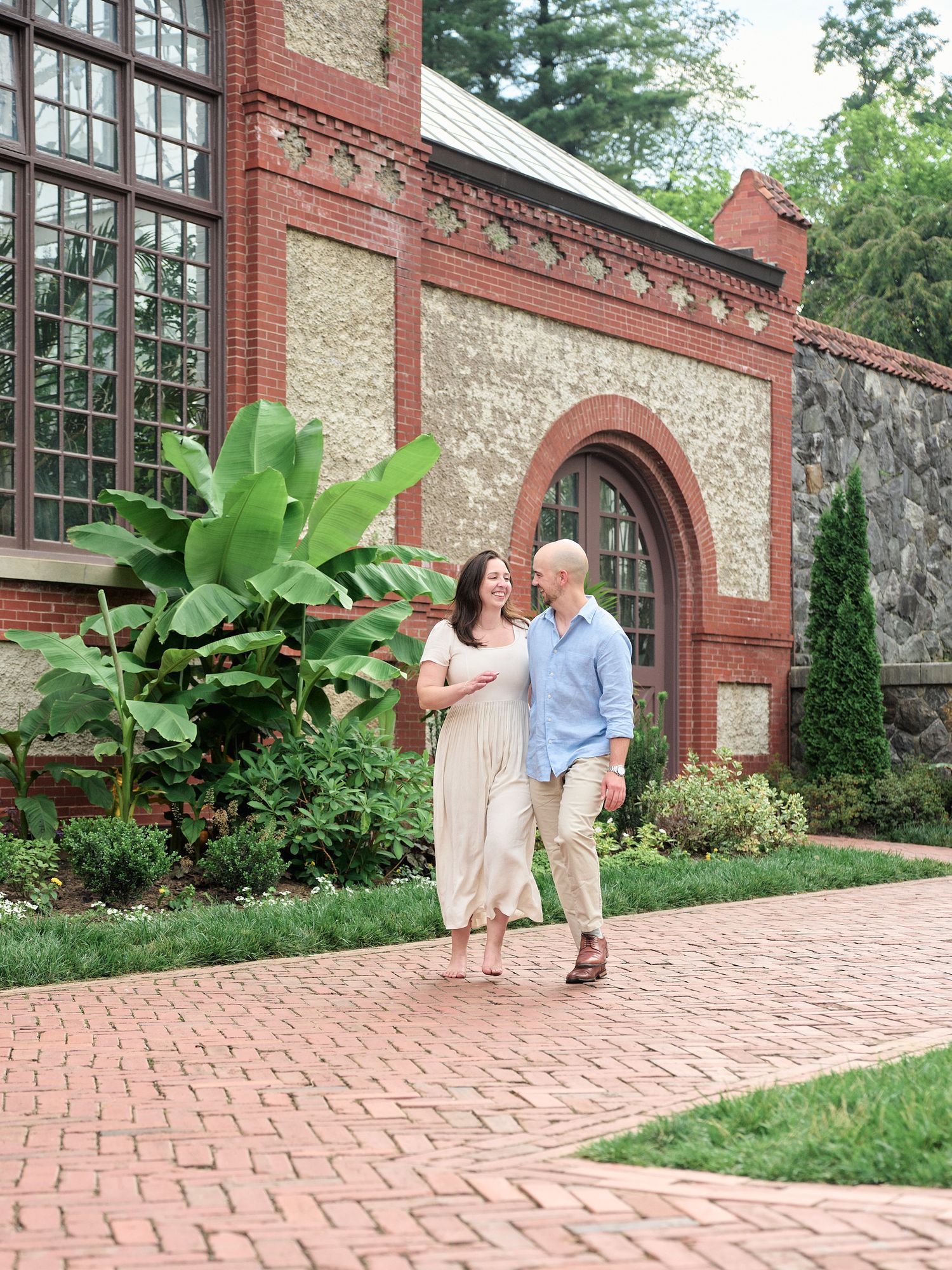 A couple walks together along a brick path beside a historic conservatory building.
