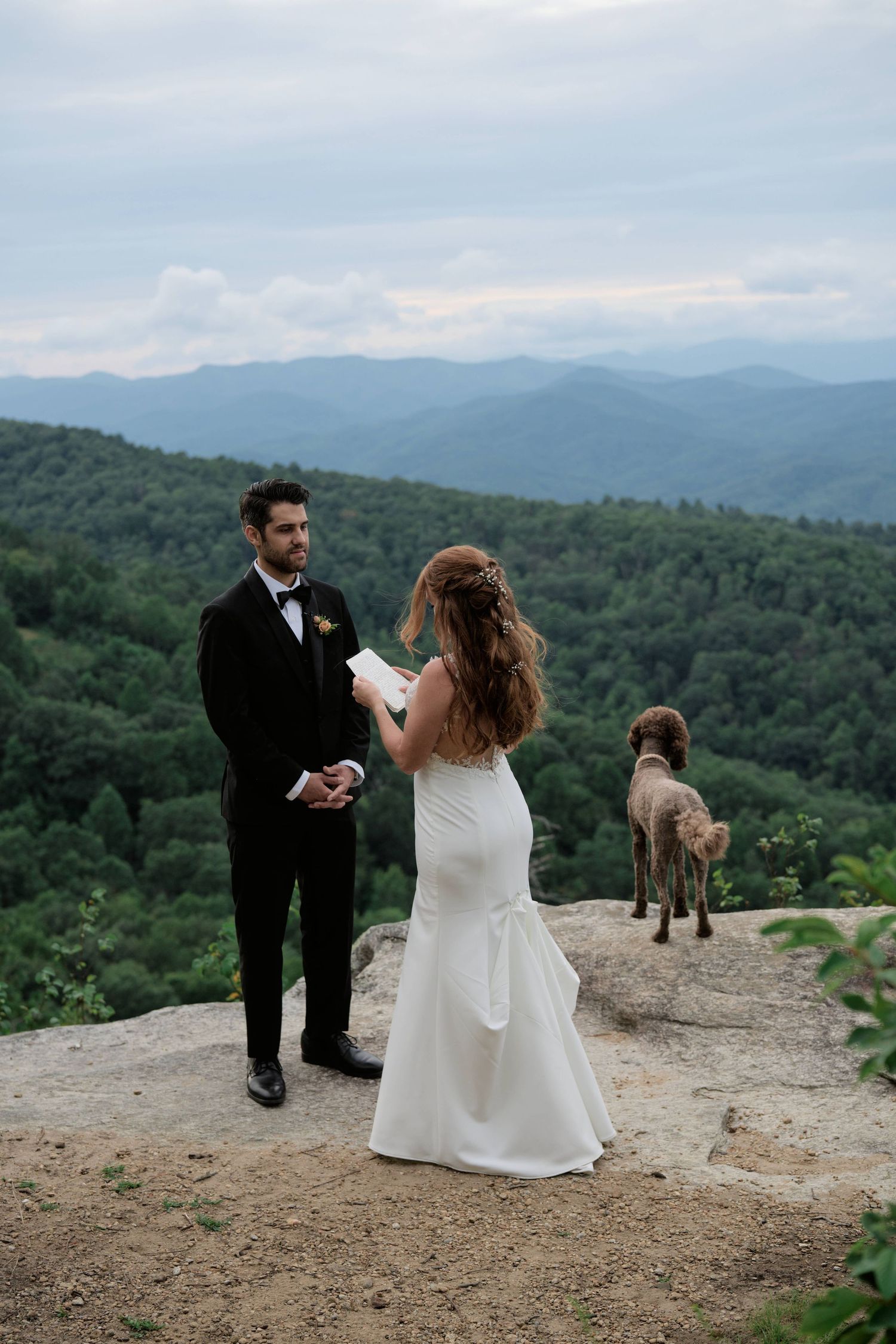 A couple in wedding attire stands at a scenic mountain overlook with rolling hills and forests stretching into the distance.
