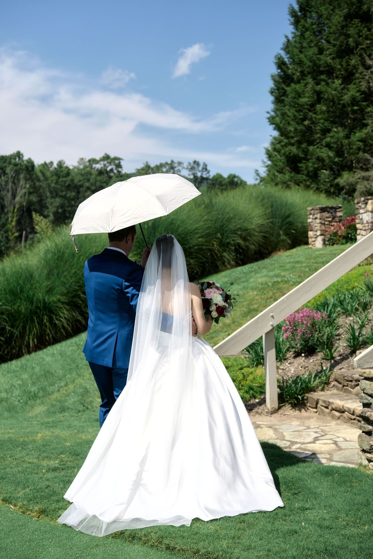 A couple under a white umbrella on a landscaped garden path during their wedding day.