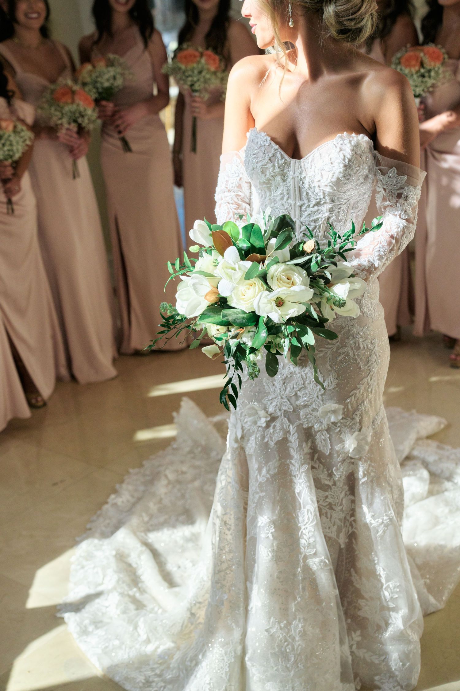A detailed view of a lace wedding dress and white rose bouquet with bridesmaids in pink dresses.