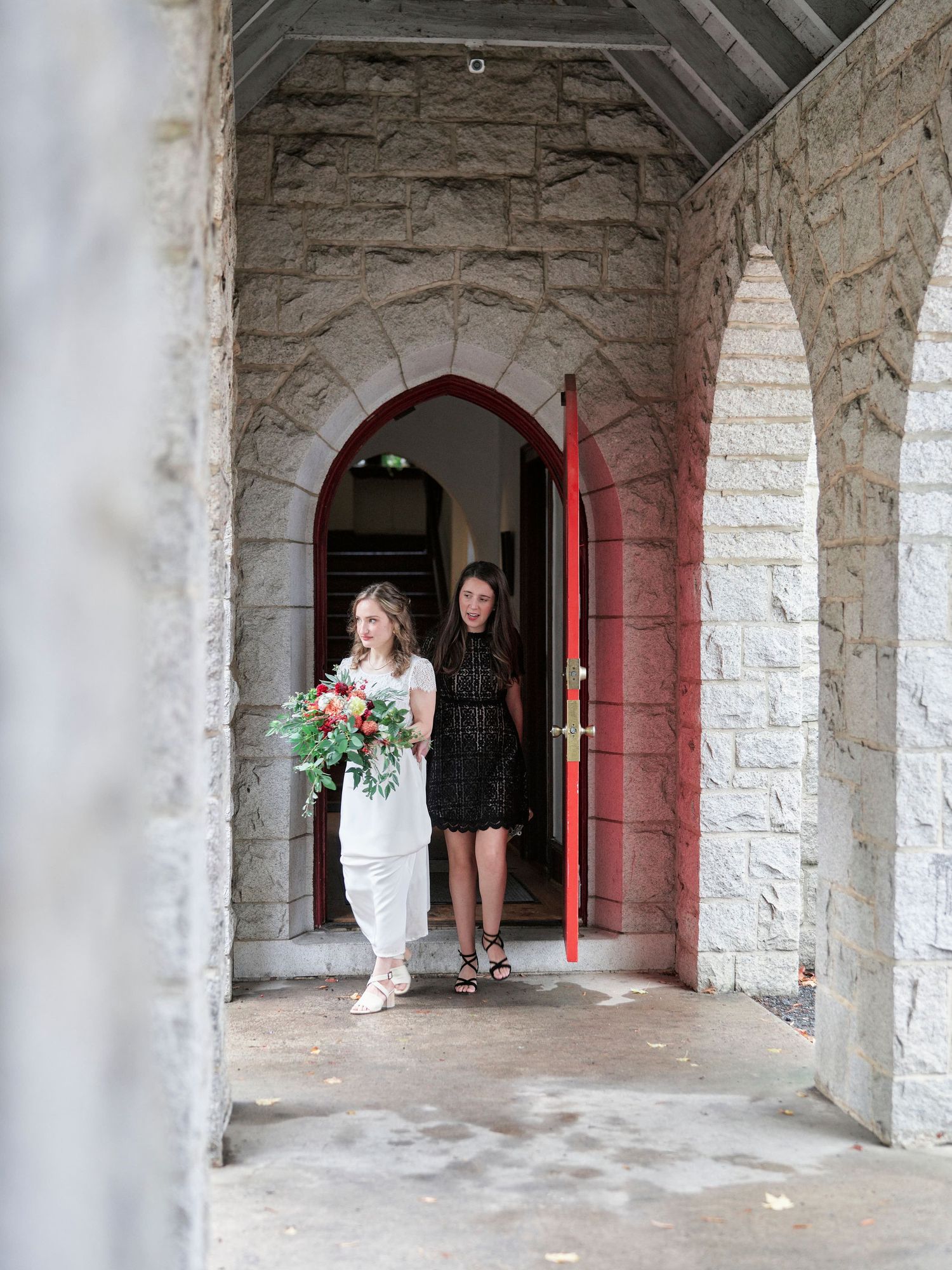 A bride carrying a colorful bouquet exits through a red church door with an attendant.
