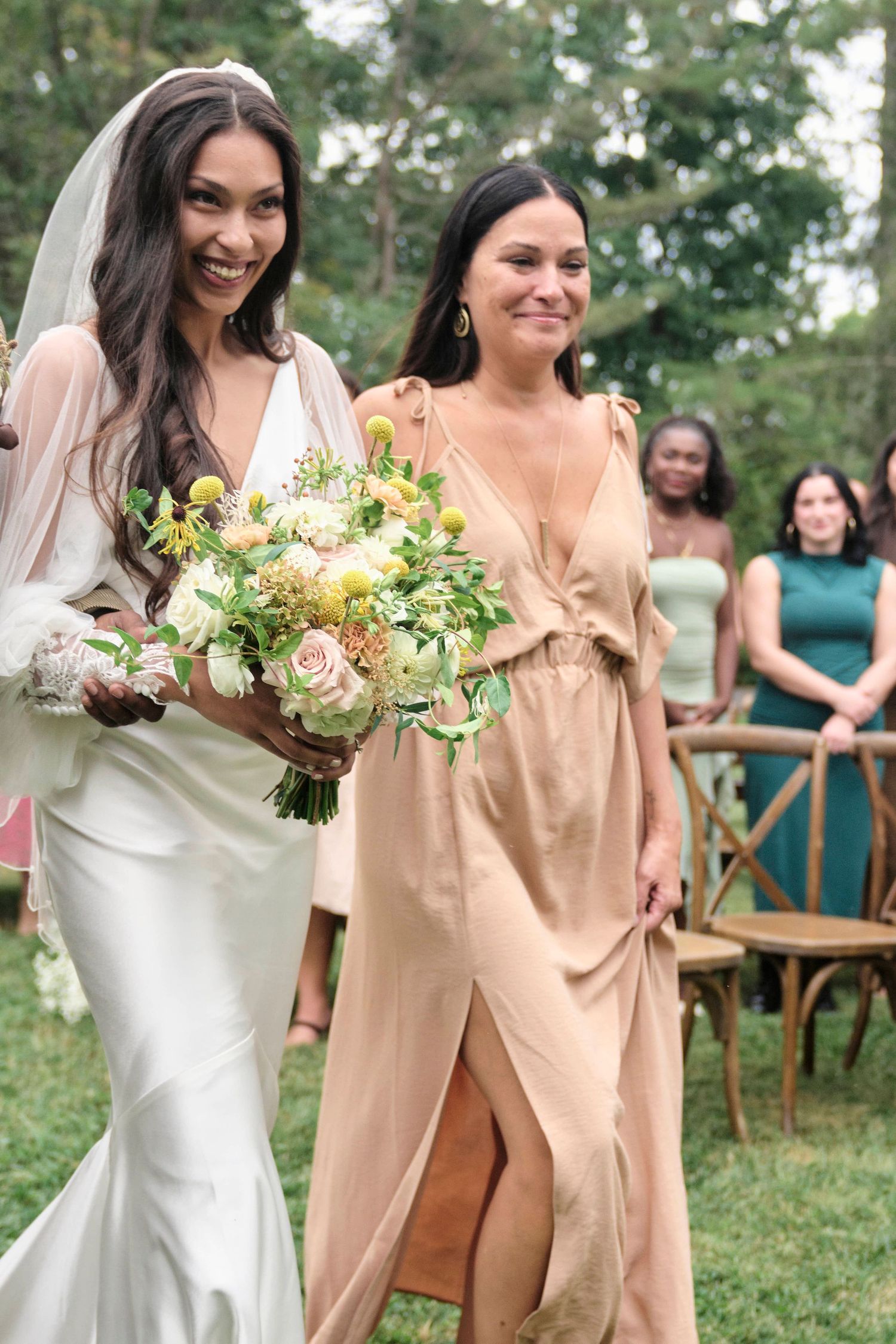 Wedding party members in white and nude-colored dresses pose with floral bouquet outdoors.