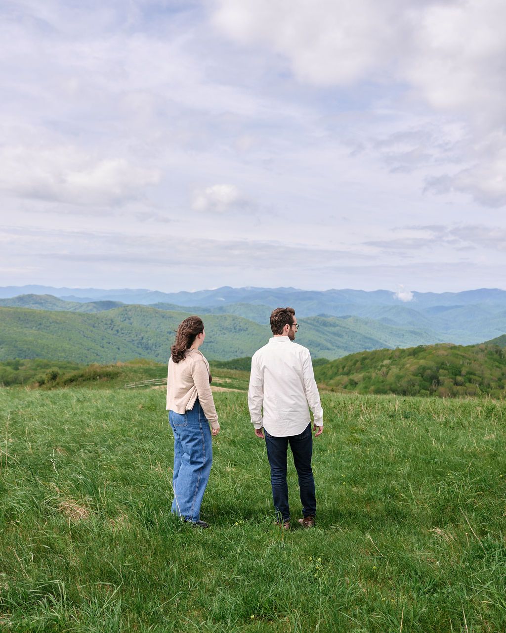 Two people stand together in a vast green mountain meadow with rolling hills and cloudy skies in the background.