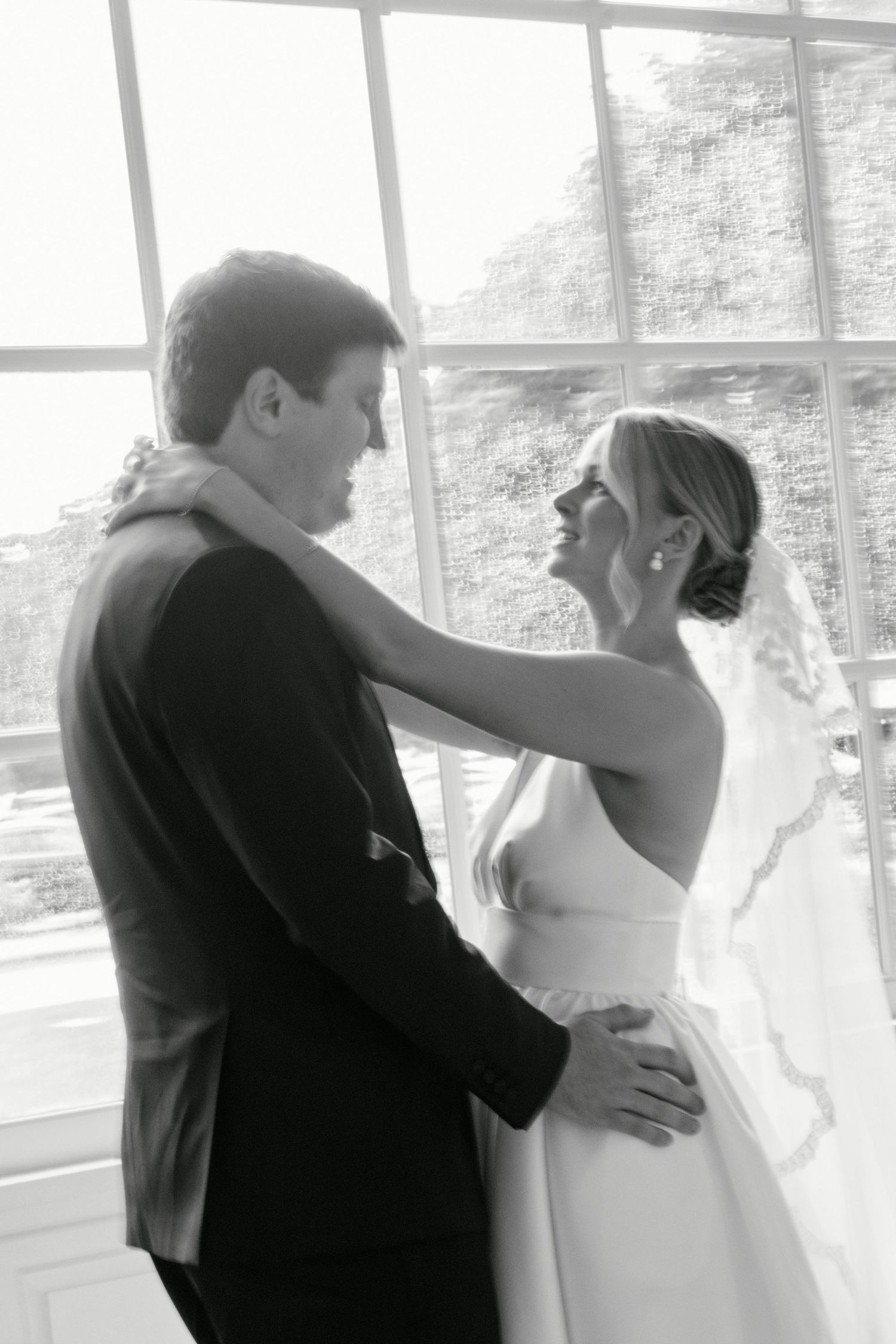 Black and white portrait captures tender moment between newlyweds by window.