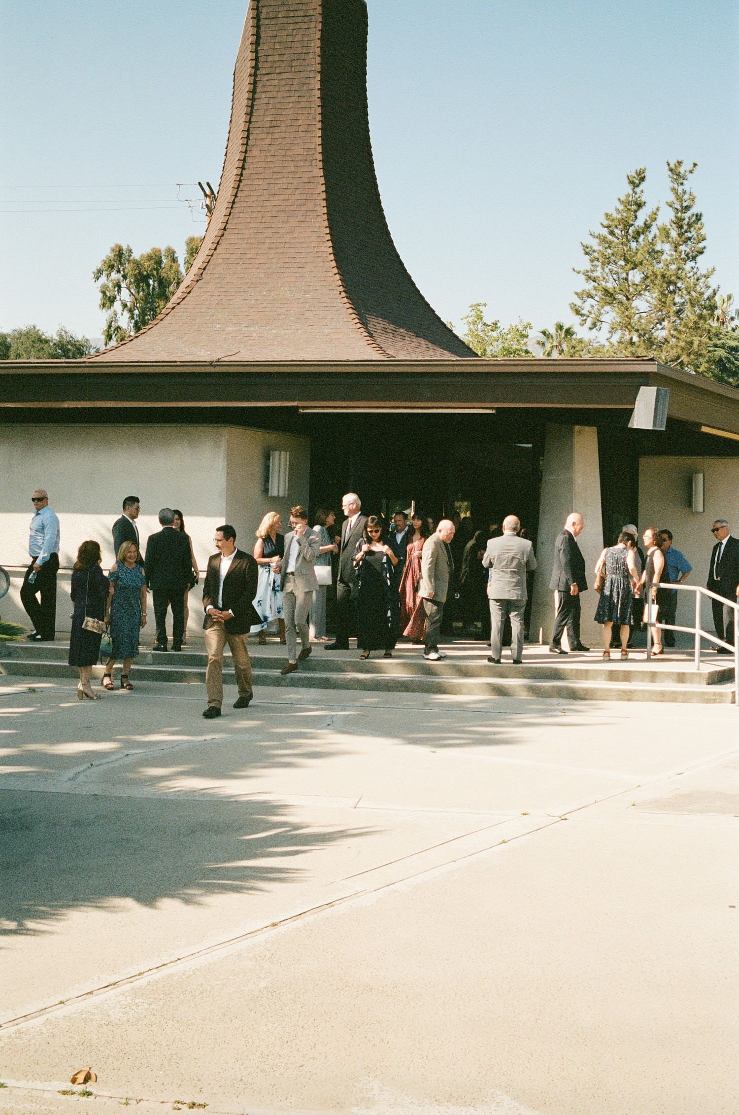 People queuing outside building with distinctive curved roof on sunny day.