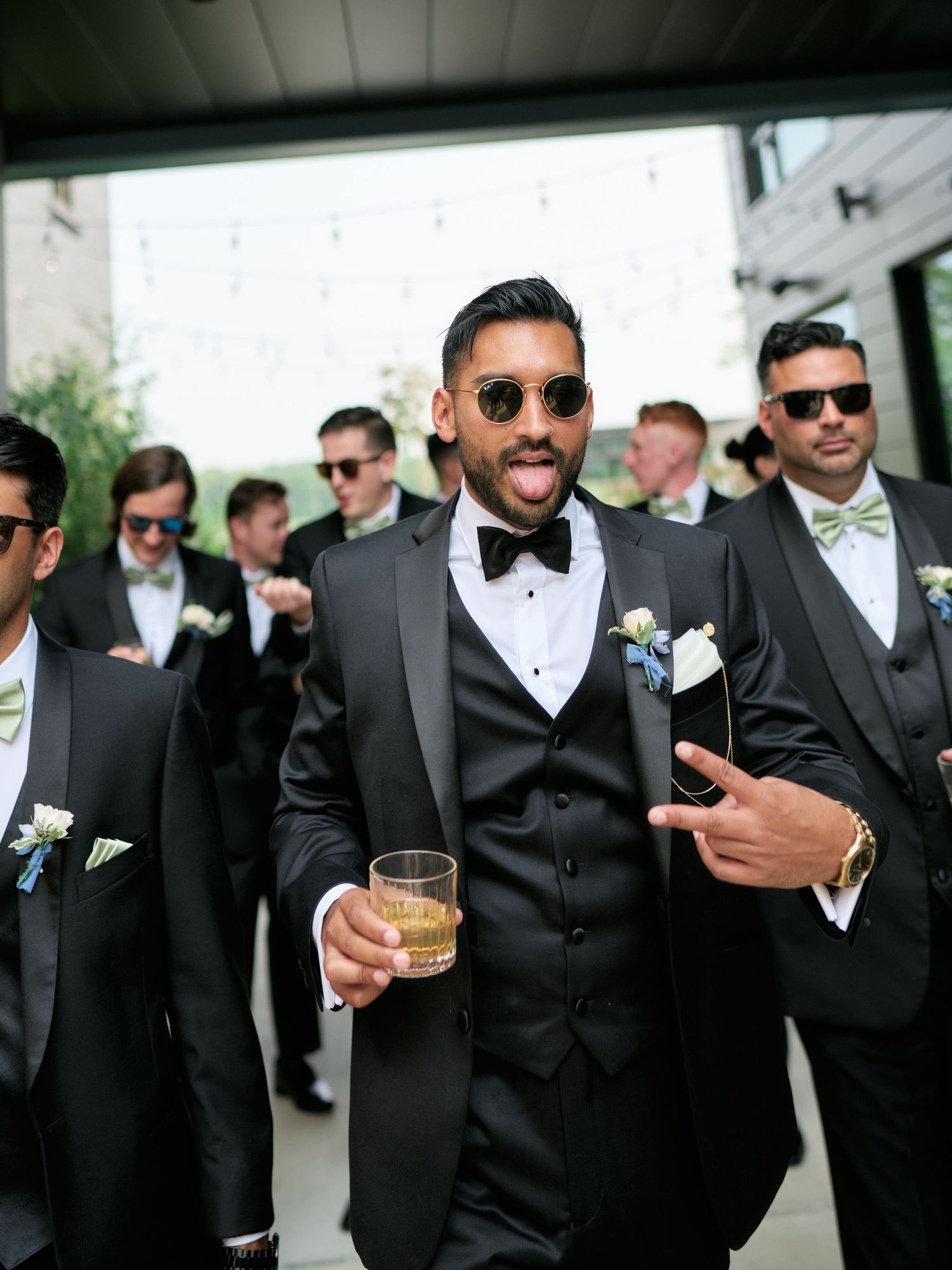 A group of groomsmen in black tuxedos and sunglasses share drinks and smiles during a wedding celebration.