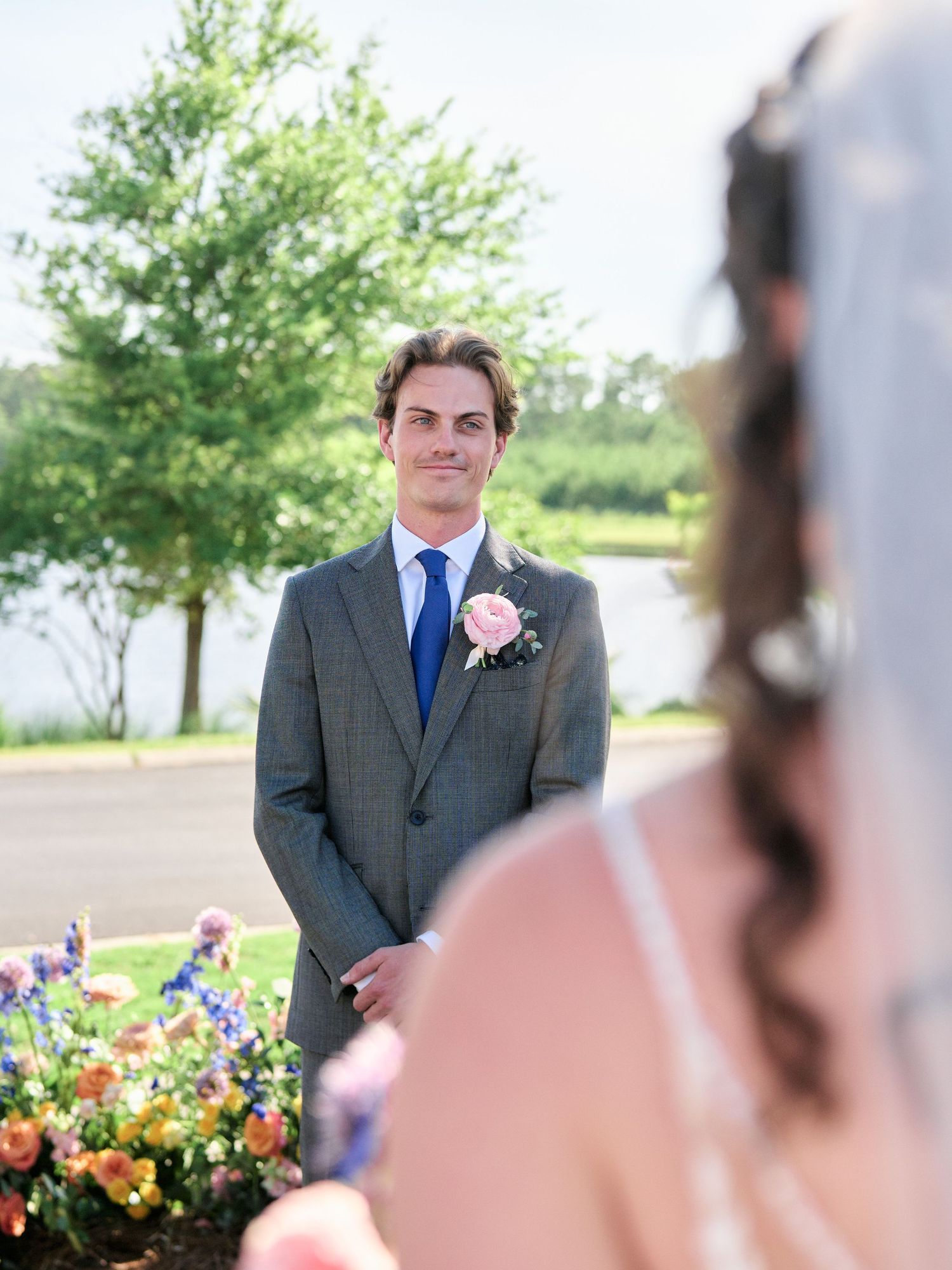 A groom in a grey suit with pink boutonniere stands outdoors during a wedding ceremony with flowers and trees in background.