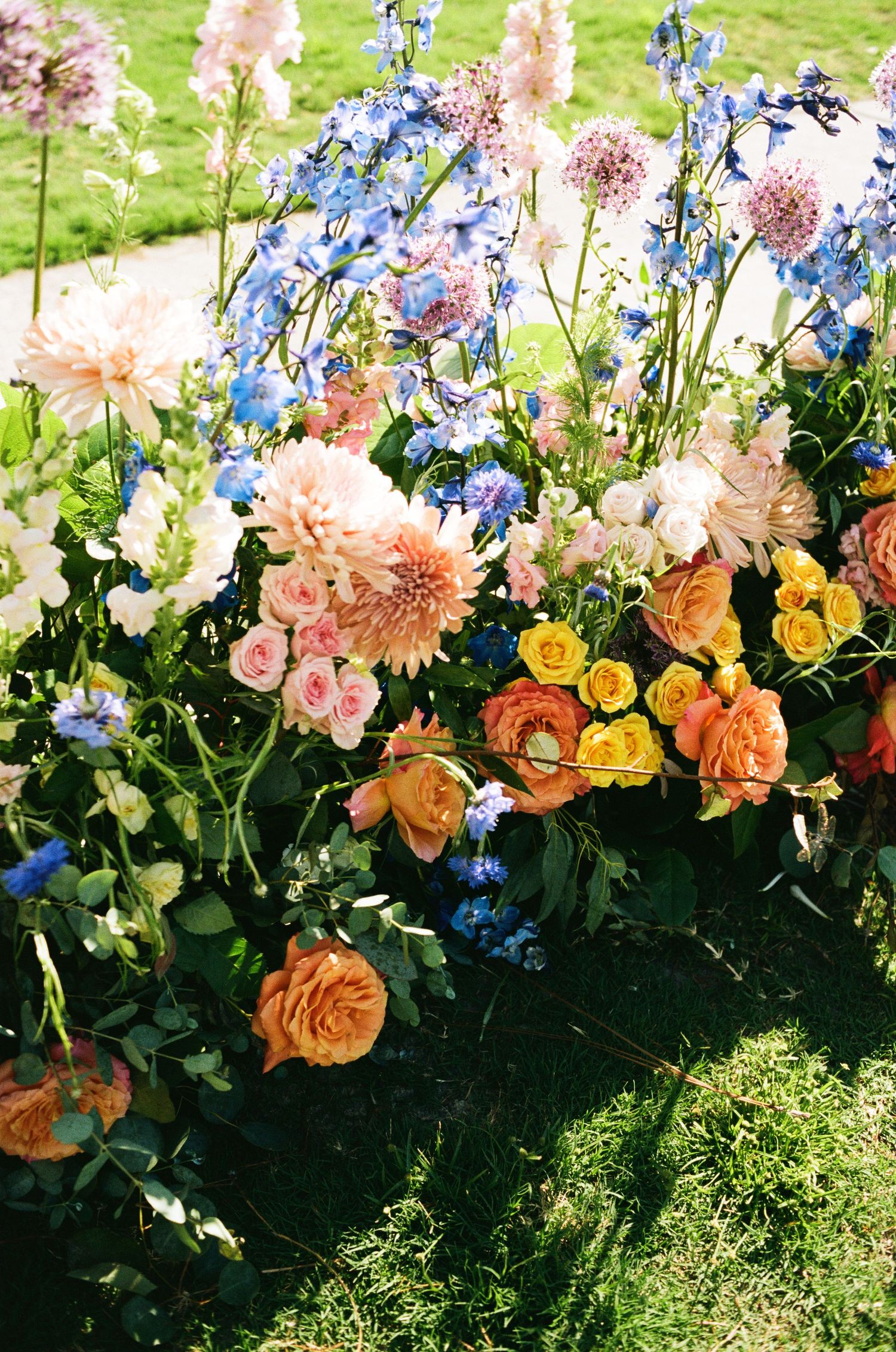 Colorful garden flowers including peach roses, blue delphiniums, and pink dahlias arranged in a lush outdoor display.