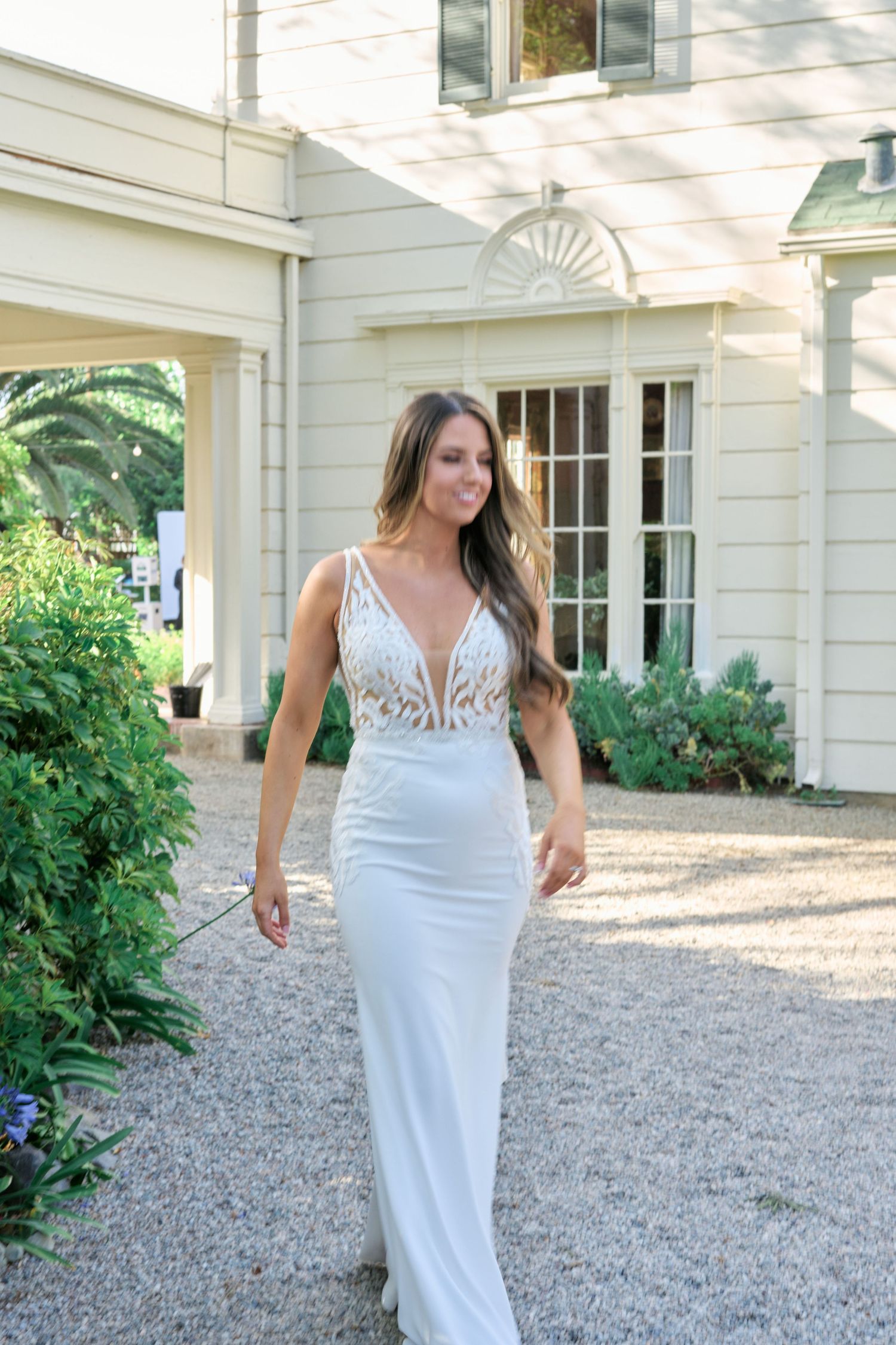 A white fitted wedding gown with delicate lace details and plunging neckline in a garden setting.