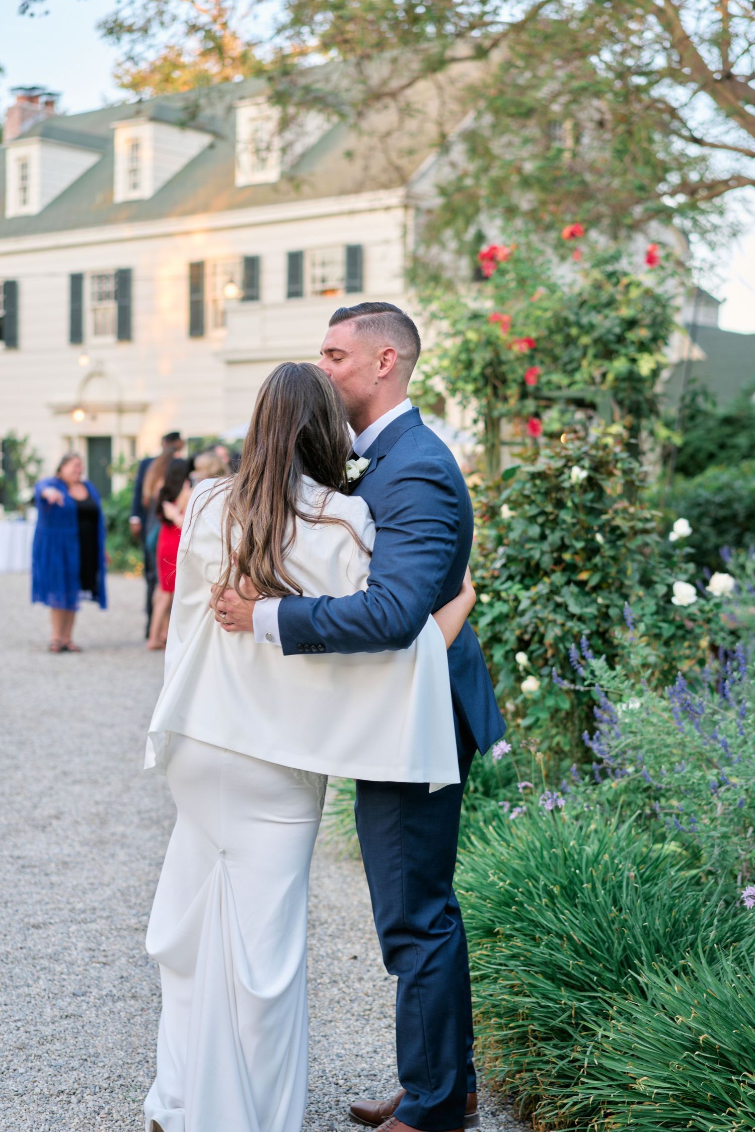 A couple embraces near a colonial-style building with flowering gardens in the background.