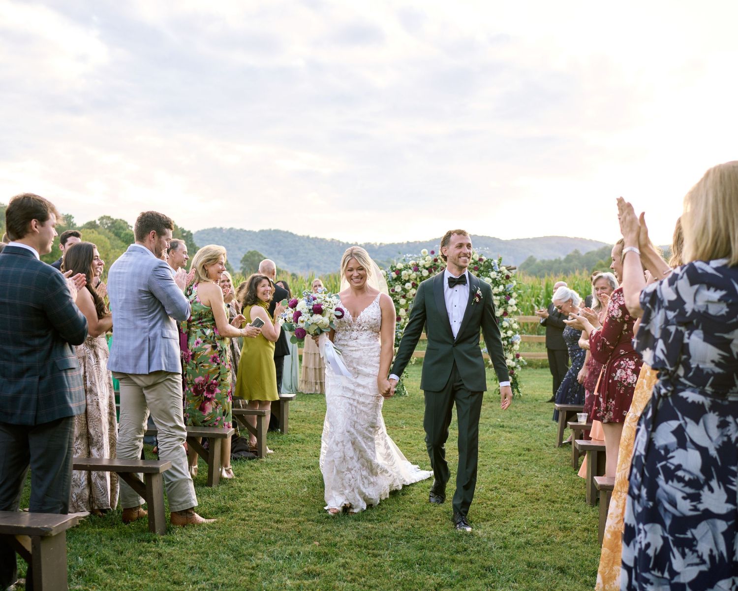 Wedding guests mingle and celebrate on a grassy hillside during an outdoor reception with mountain views in the background.