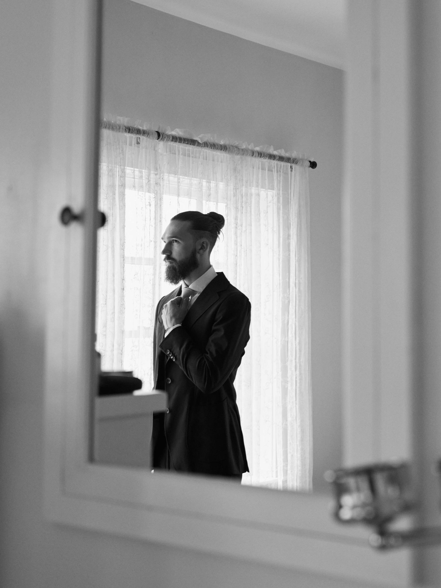 Black and white portrait of person in formal attire adjusting tie in mirror.