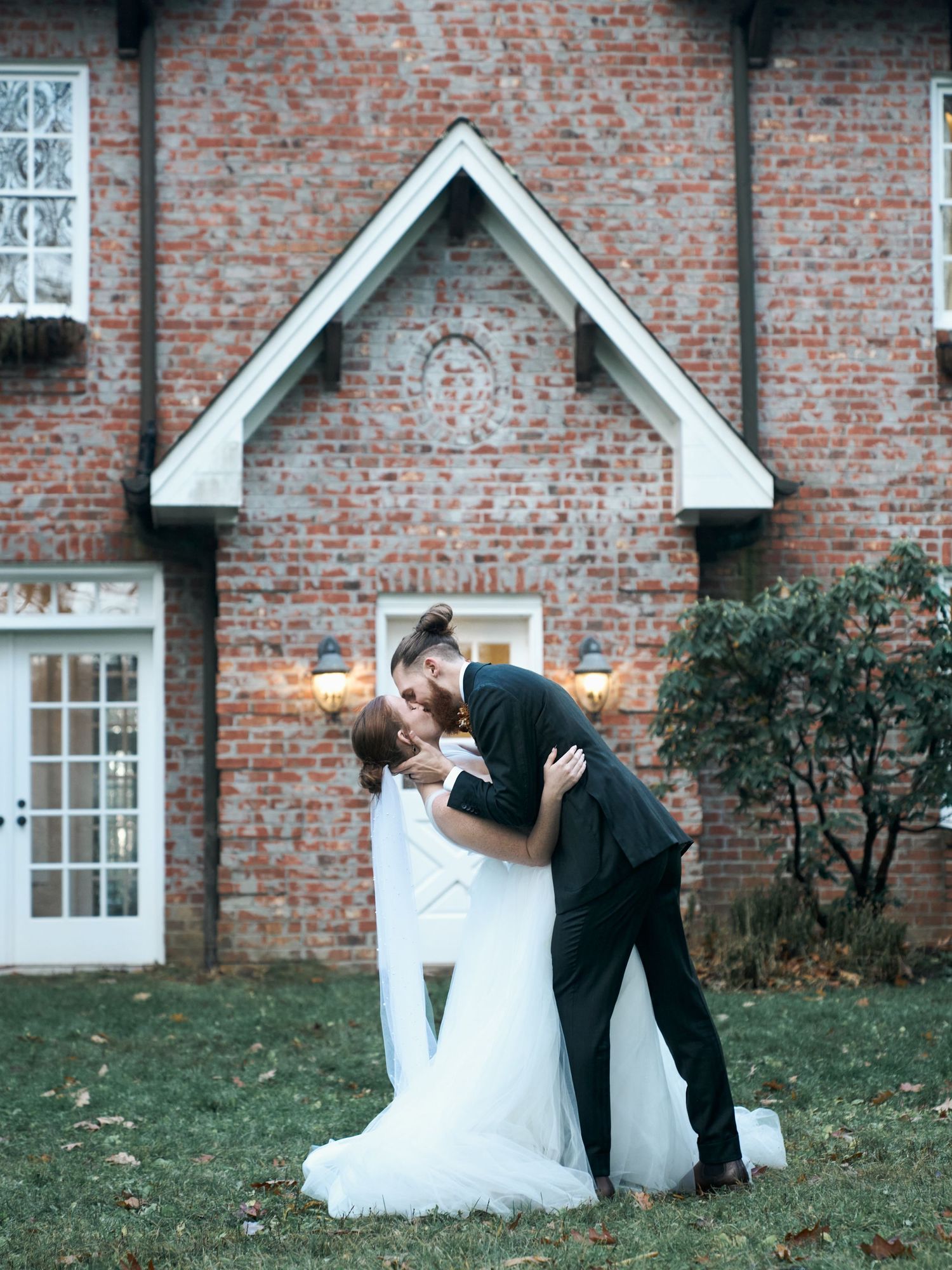 A romantic wedding pose in front of a brick house with white trim and windows.