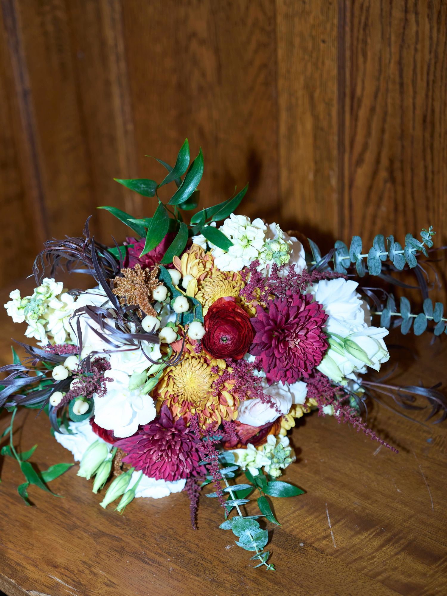 Vibrant wedding bouquet featuring burgundy dahlias, eucalyptus, and white flowers on wooden surface.