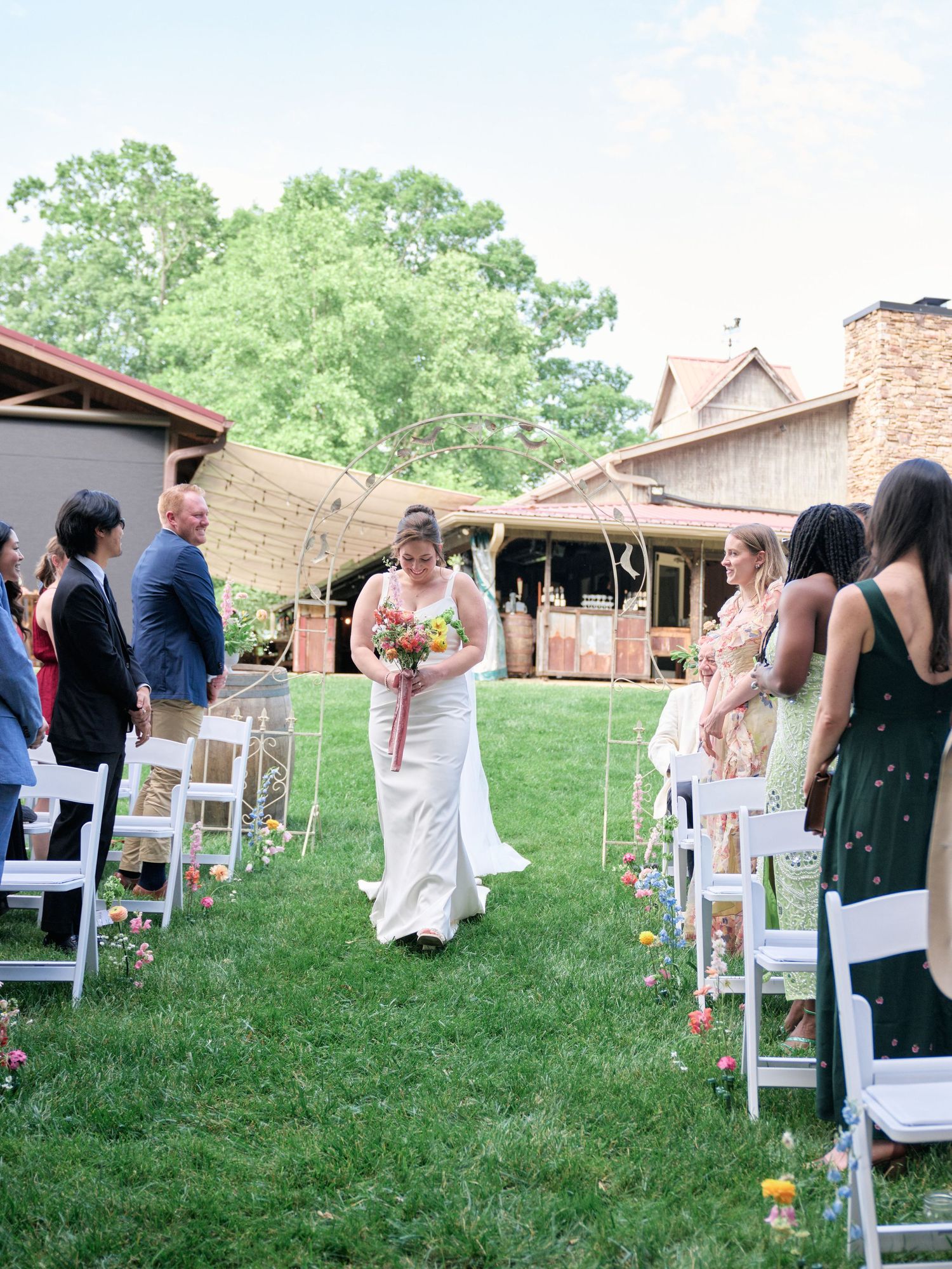 Wedding ceremony procession on grass pathway lined with white chairs and lanterns.