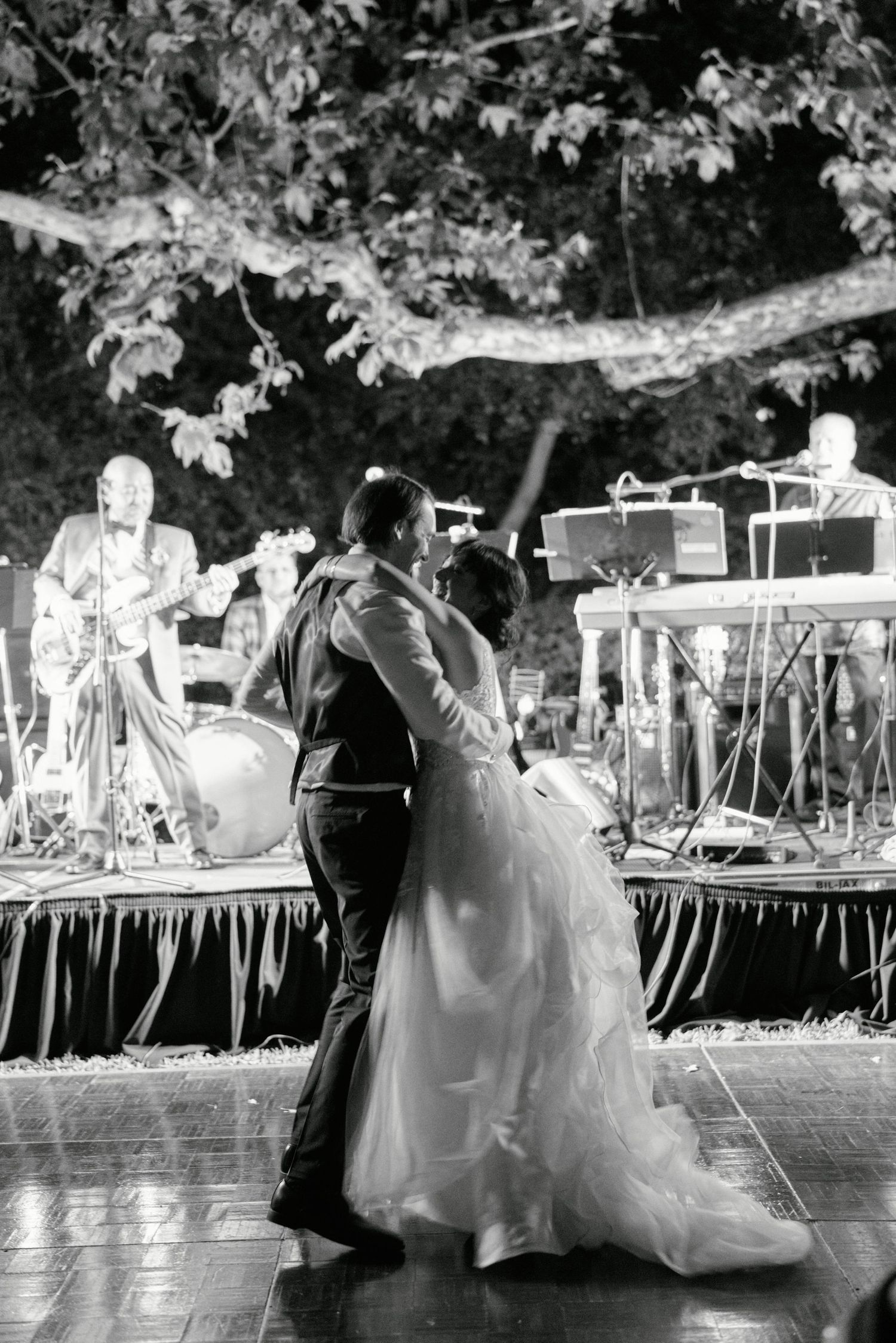 A couple dances at their outdoor wedding reception with a band performing on a decorated stage.