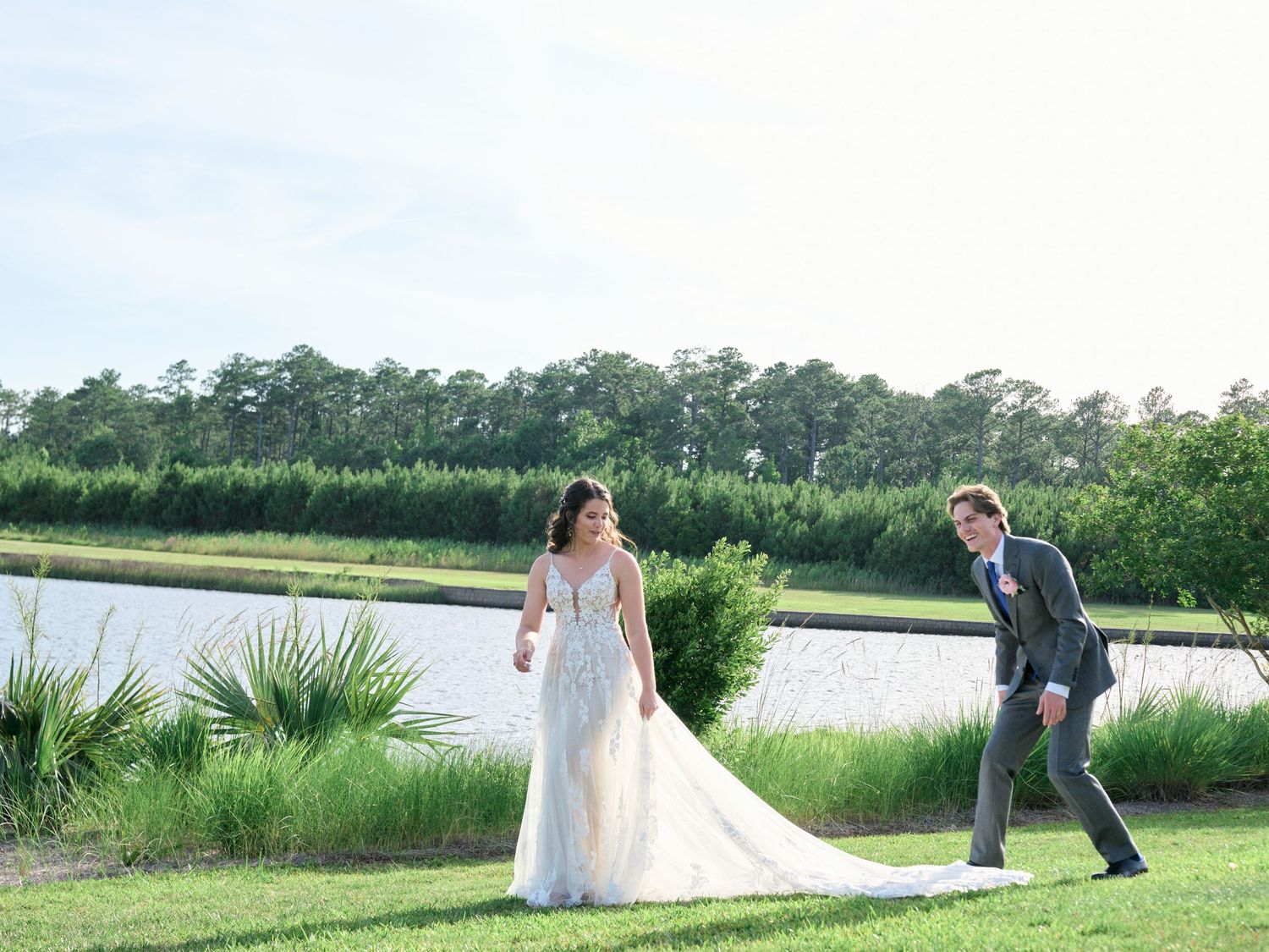 A playful wedding couple poses by a serene lake surrounded by lush greenery and marsh grass.