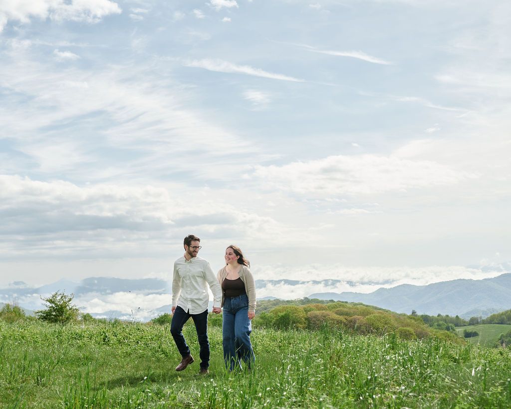 Two people walking together in a green meadow with mountains and cloudy skies in the background.