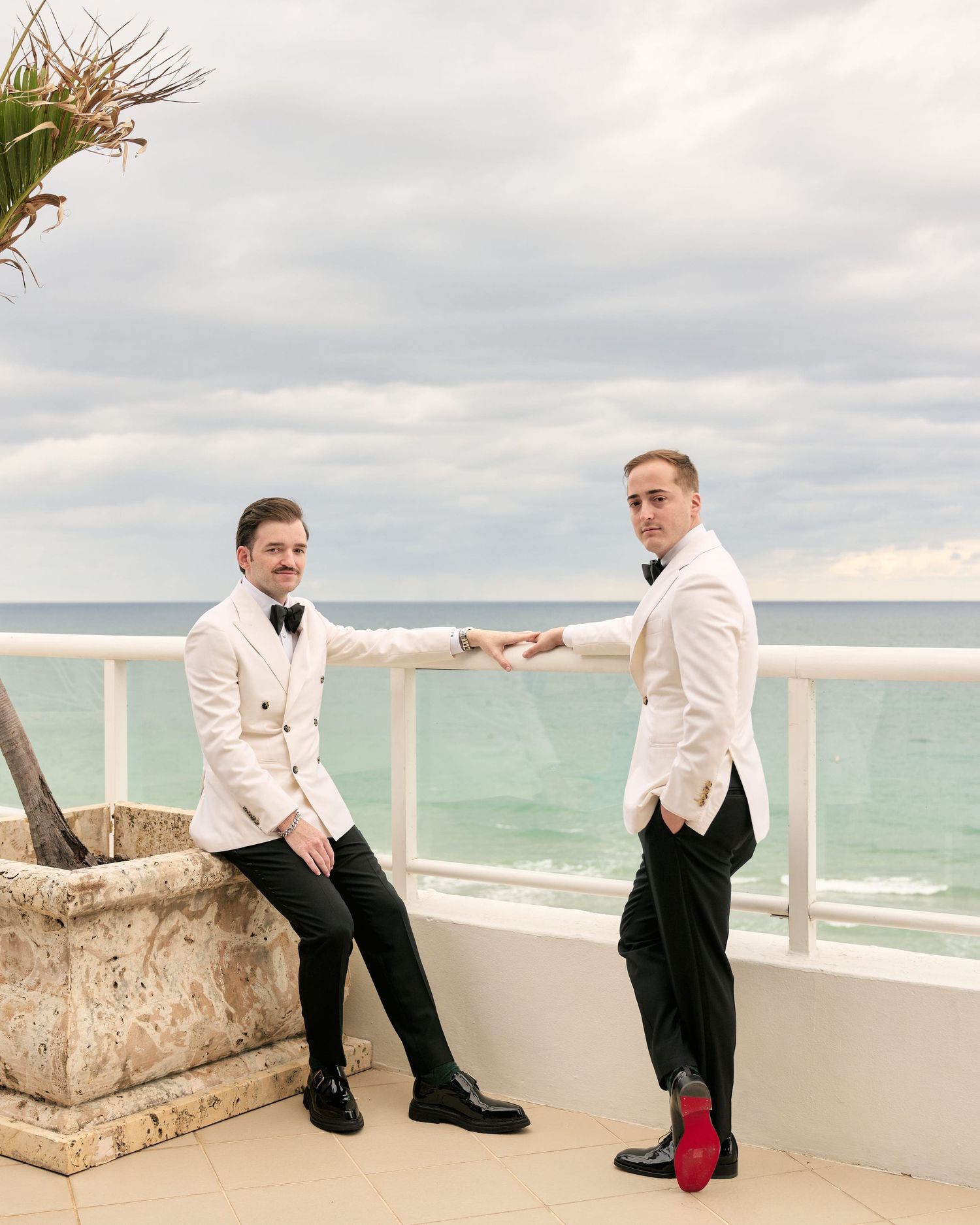 Two people in white dinner jackets pose on a balcony overlooking the ocean.