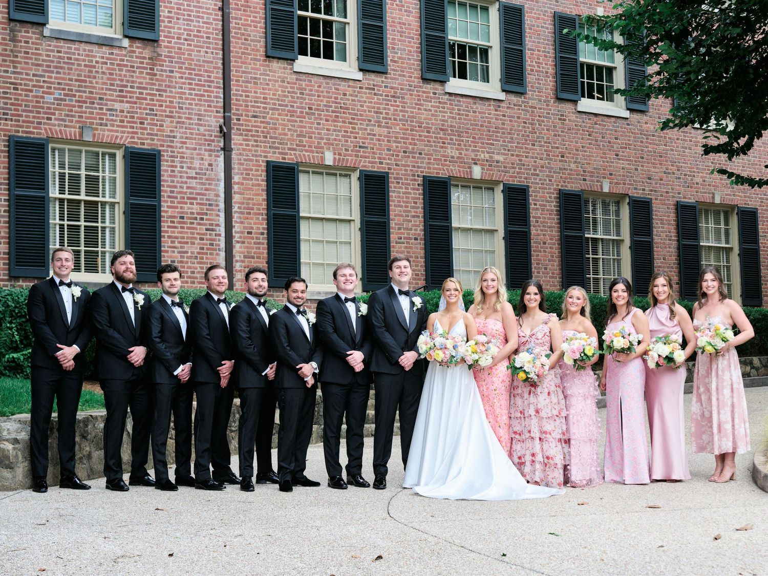 Wedding party in pink dresses and black tuxedos poses together against classic brick building backdrop.