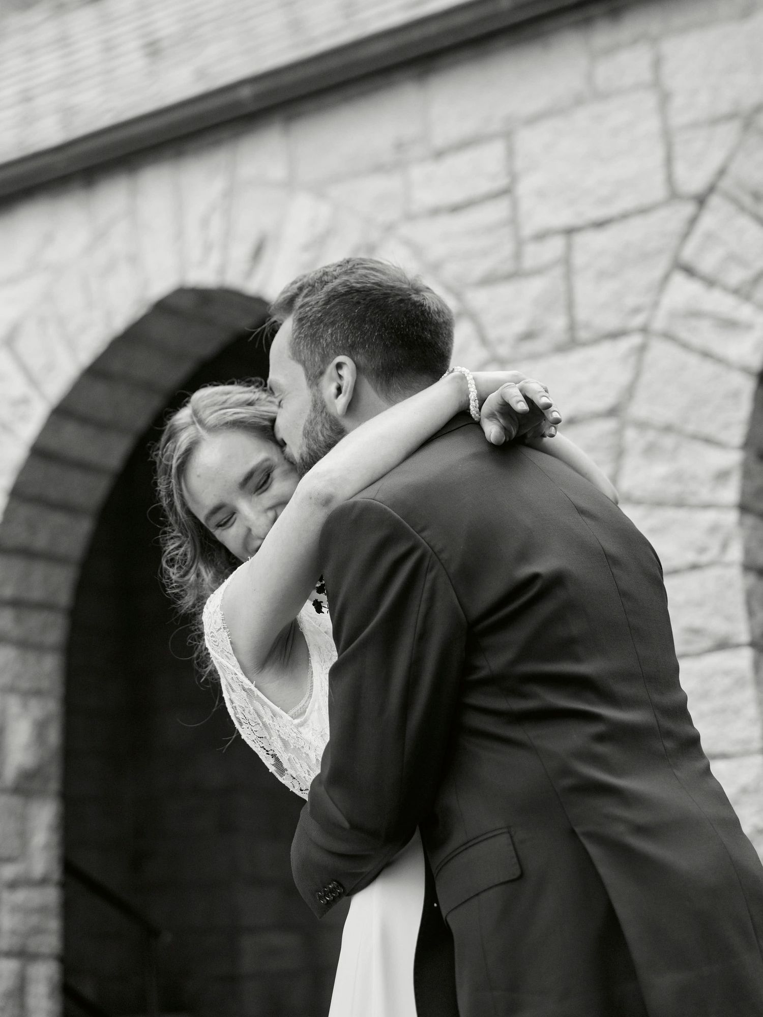 An intimate black and white wedding portrait showing a tender embrace near a stone archway.