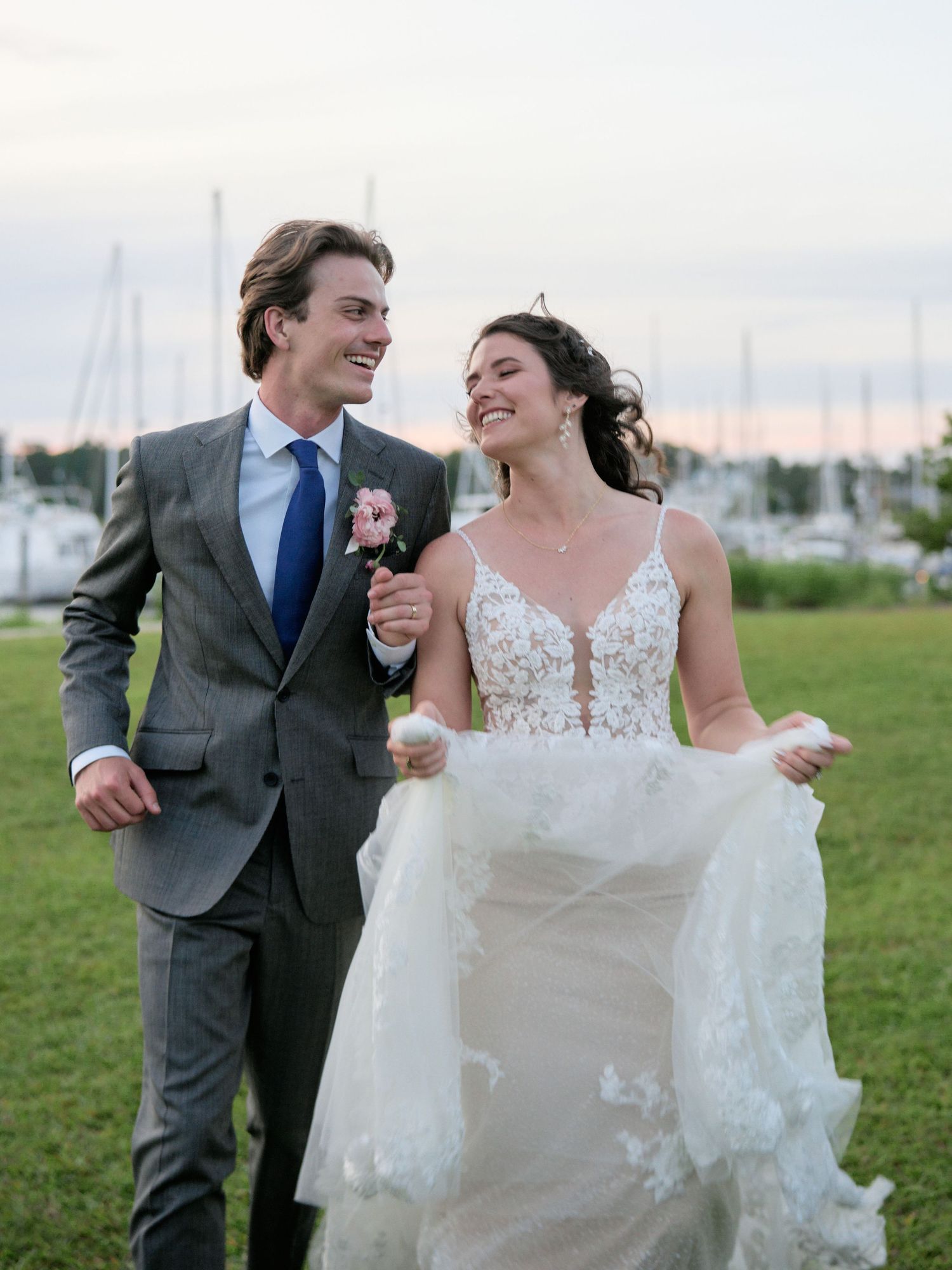A happy couple laughs together on a grassy field at sunset with a marina in the background.