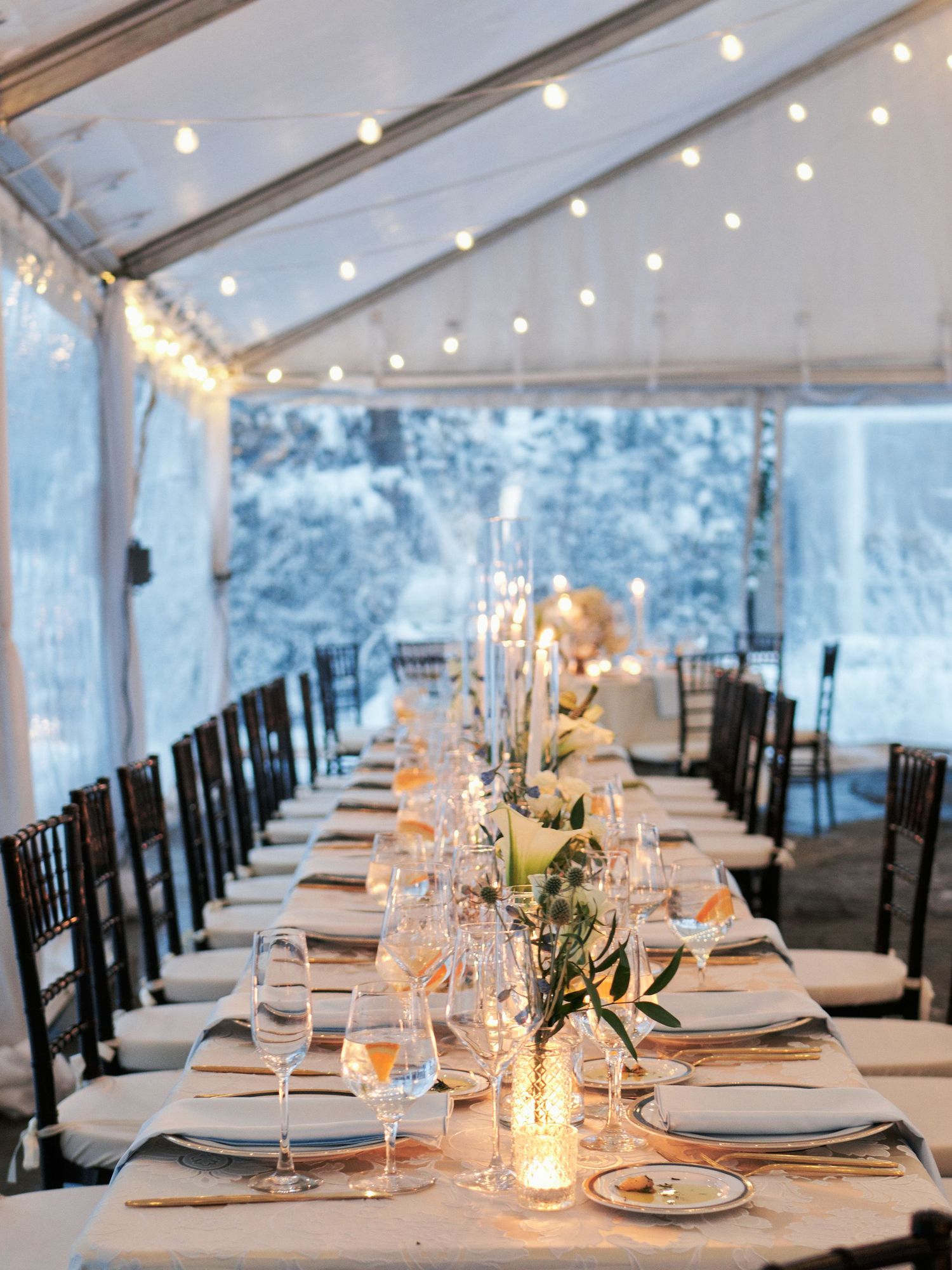 Elegant winter wedding reception under a white tent with string lights, long tables, candles, and floral centerpieces.