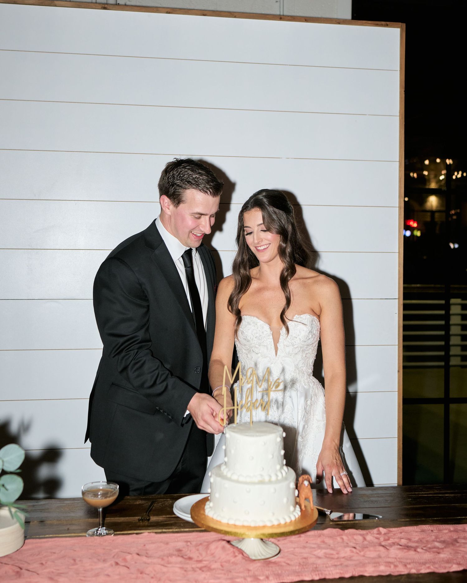 Bride and groom cutting white wedding cake together at elegant reception, groom in black suit, bride in lace gown.