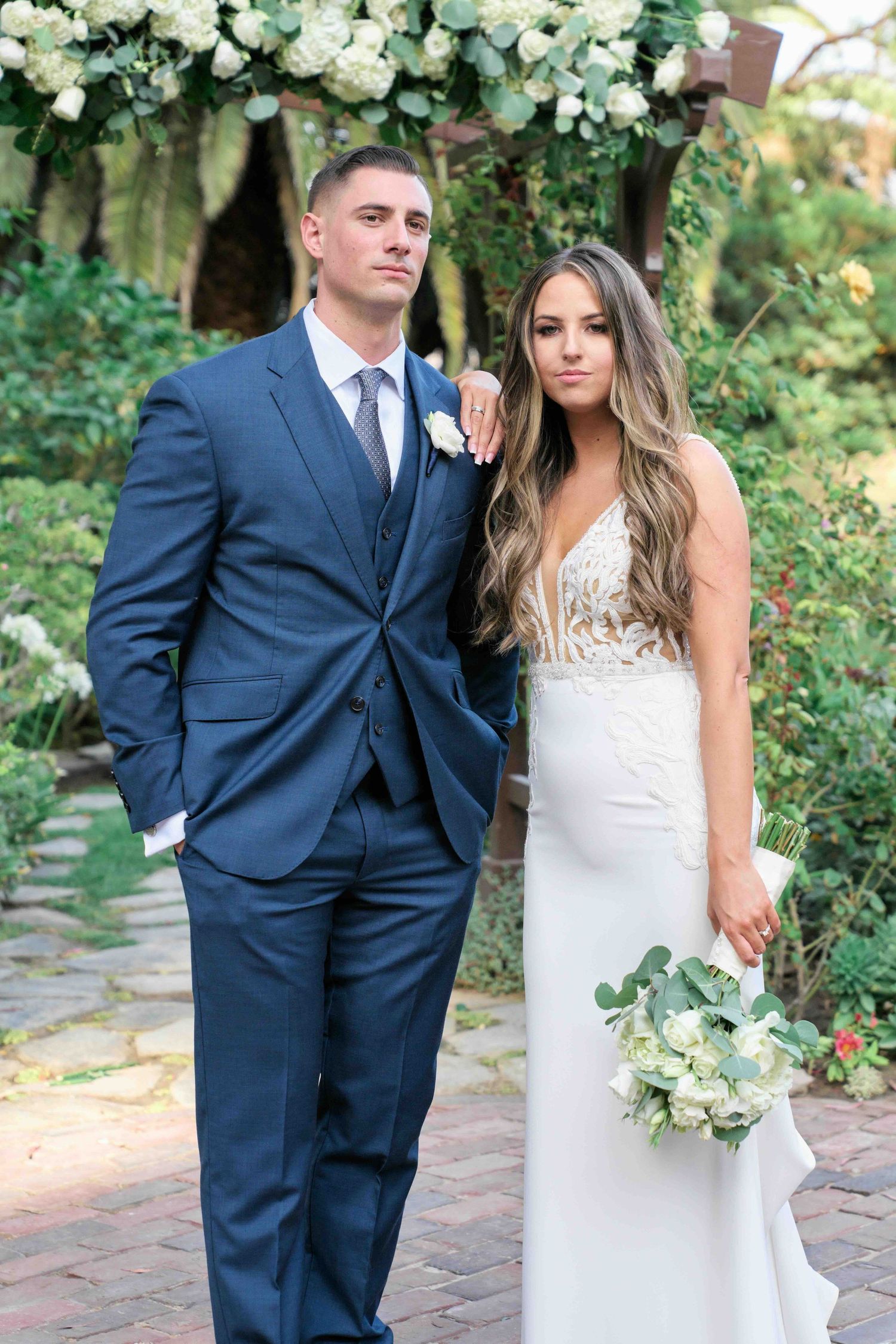 A couple in wedding attire poses together in a garden setting with white flowers and greenery.