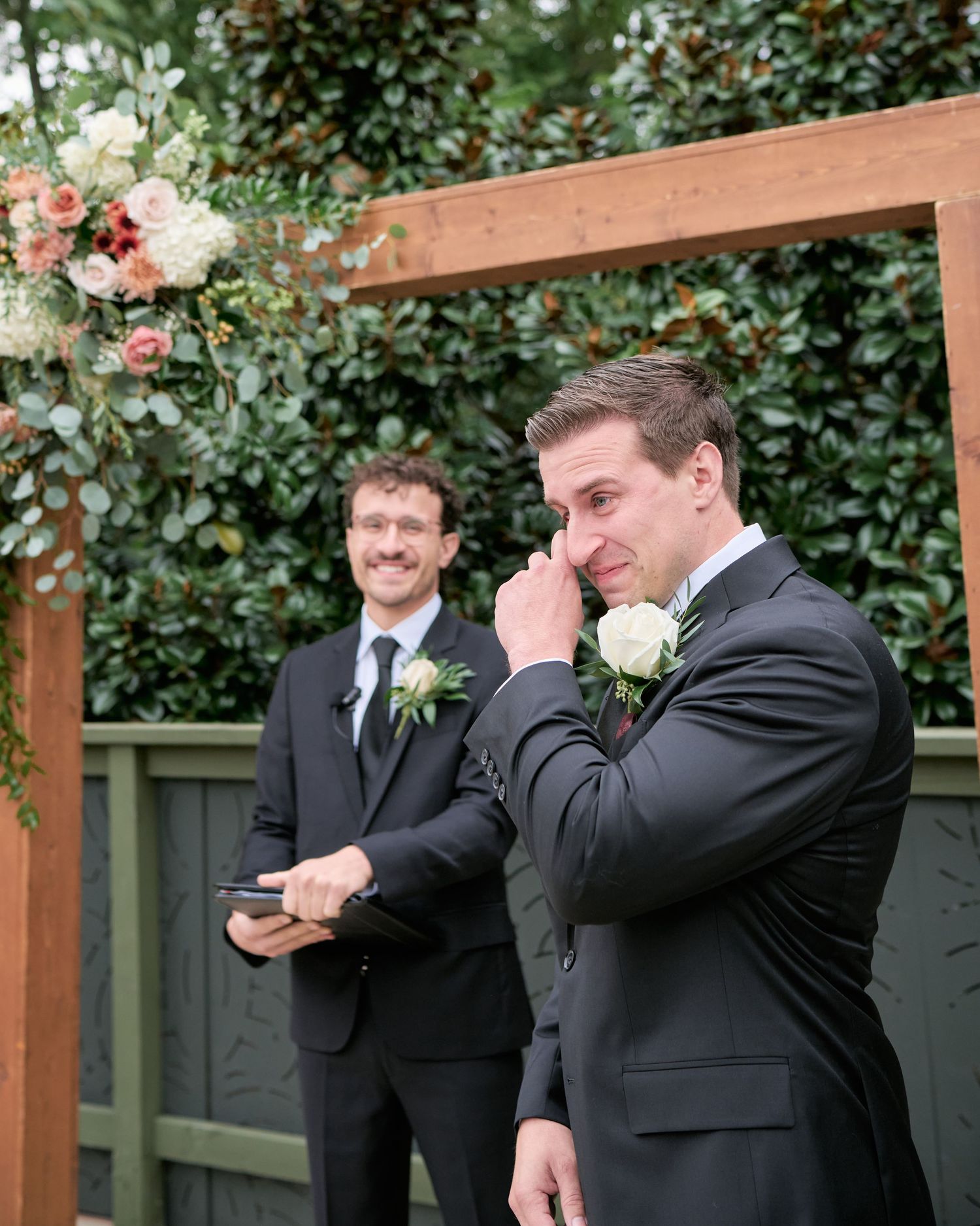 Groomsmen in black suits share a candid crying moment during a wedding session in front of greenery.