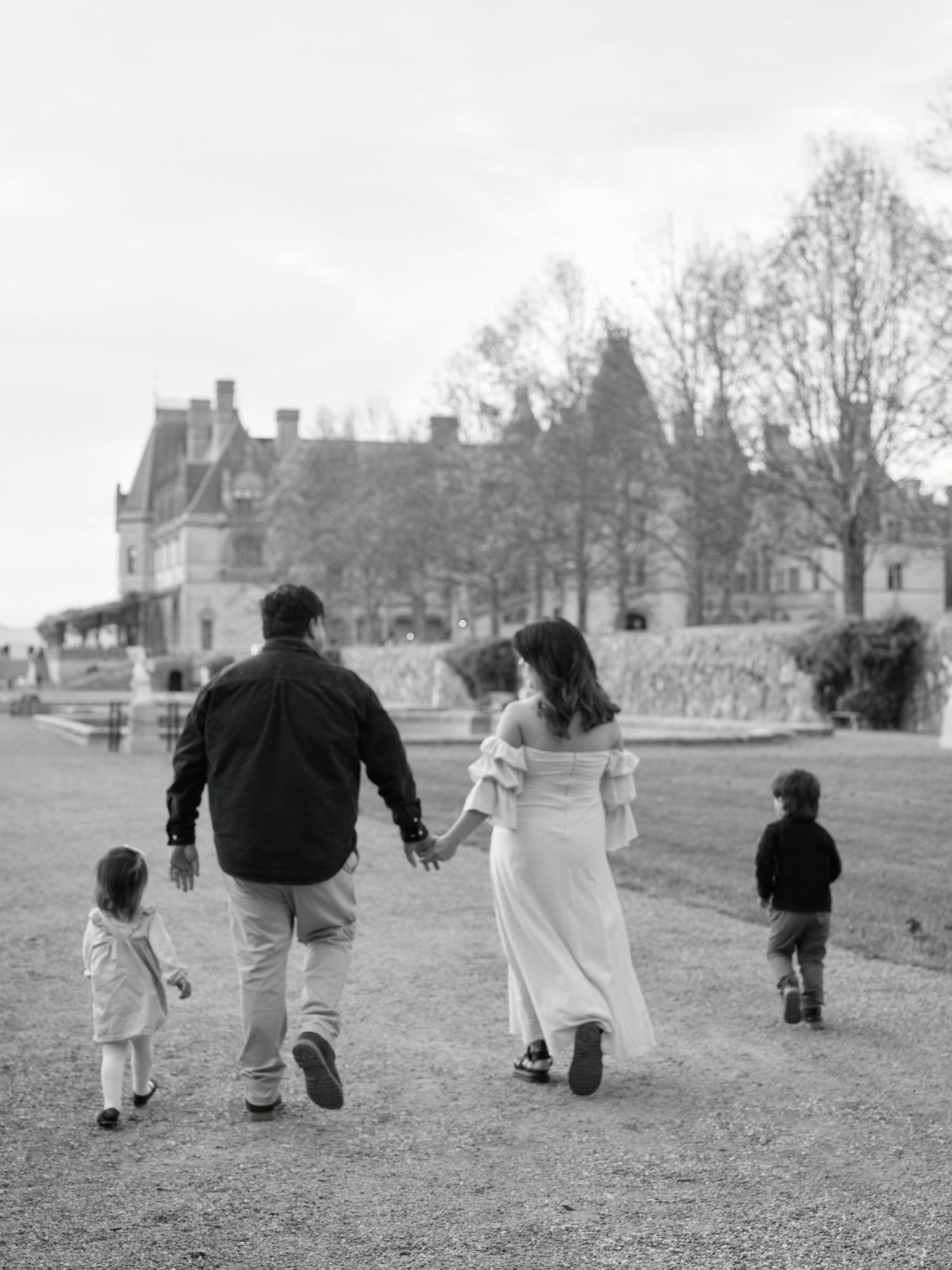 Black and white photo of a family walking toward a grand estate, parents holding hands with young children on a grassy path.