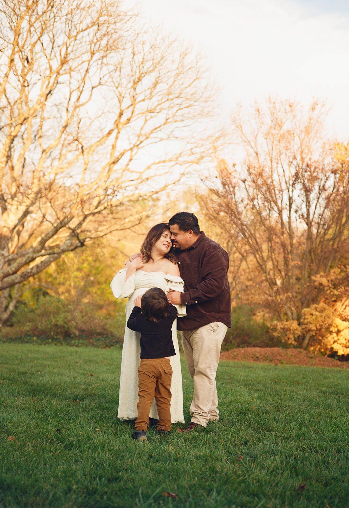 A family of three shares a tender moment outdoors during golden hour, with bare autumn trees in the background.