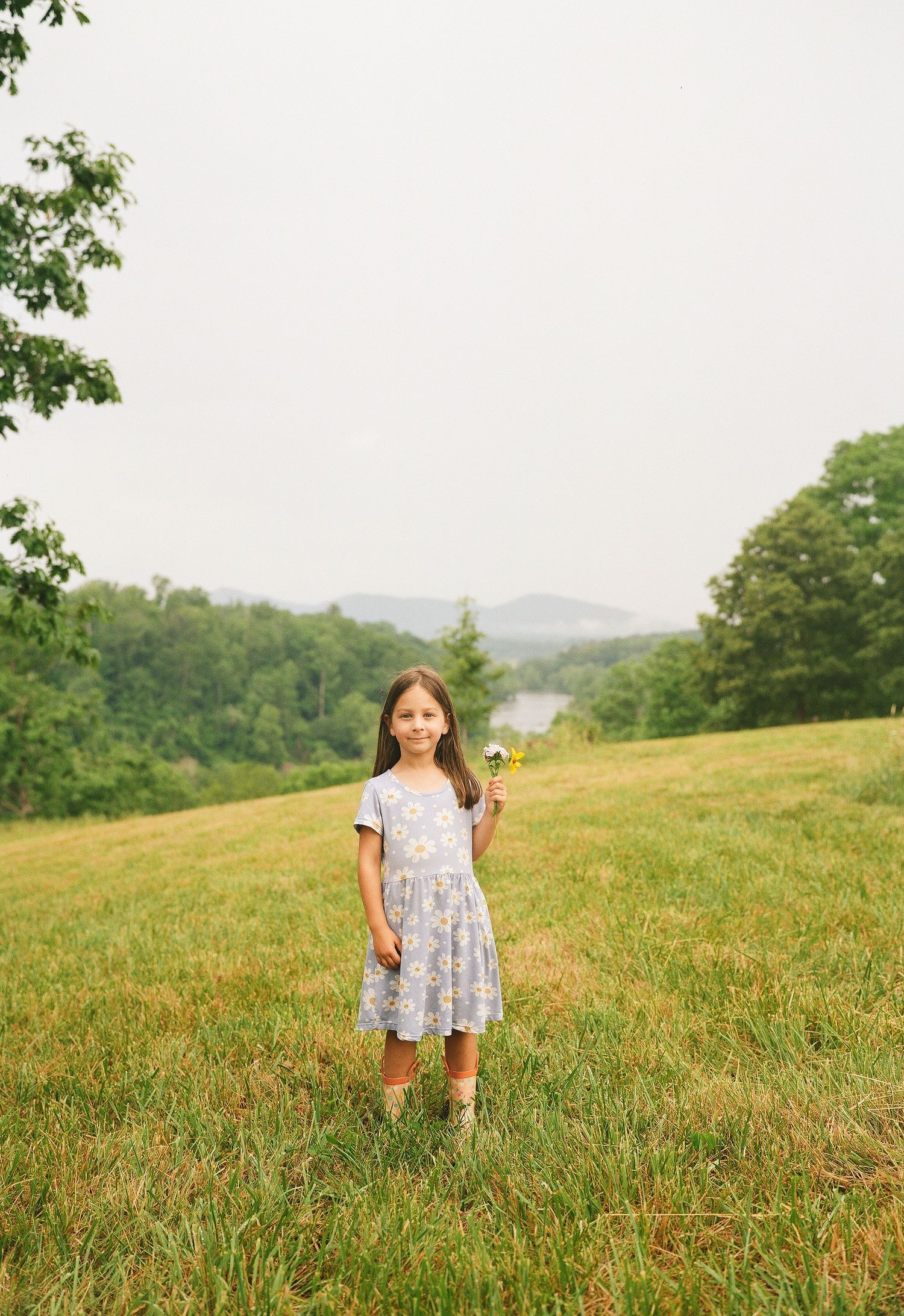 Young girl in floral dress holding wildflowers in a lush green meadow with trees and hills in the background.