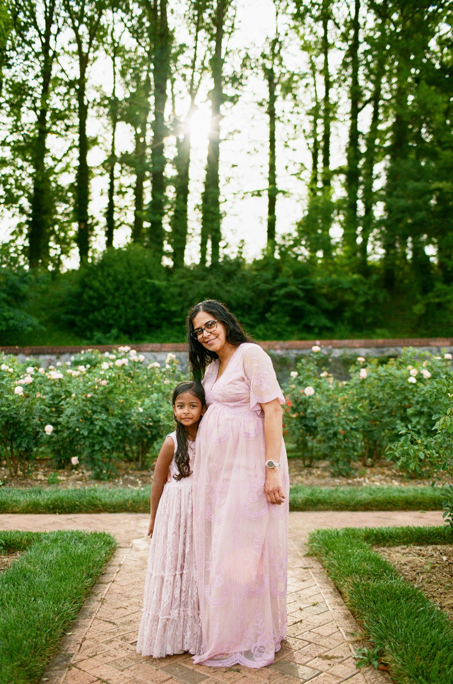 A mother and daughter in matching pink dresses pose together in a lush garden with tall trees and roses.