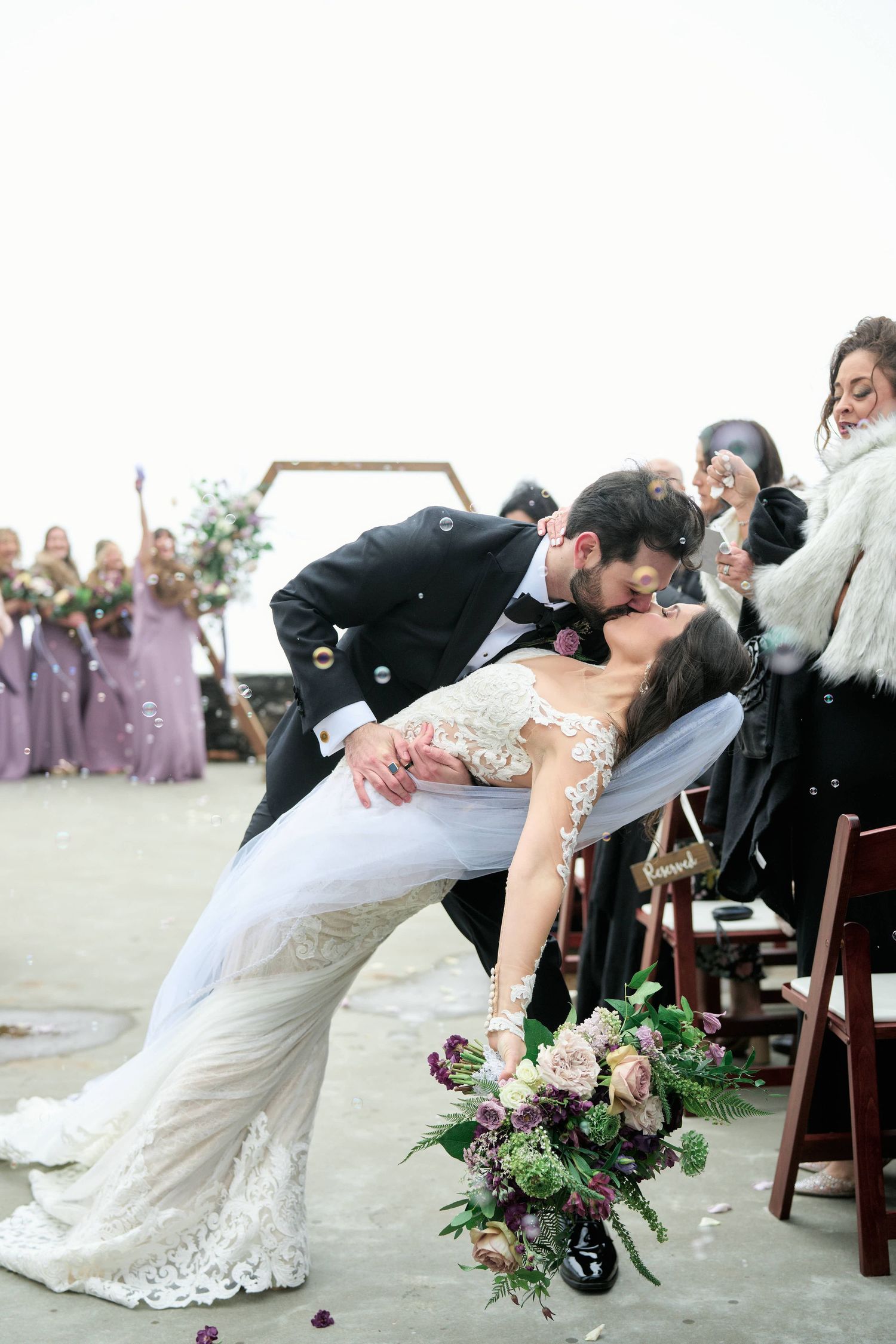 Groom dips bride for a romantic kiss at outdoor wedding ceremony with purple bridesmaid dresses and floral bouquet.