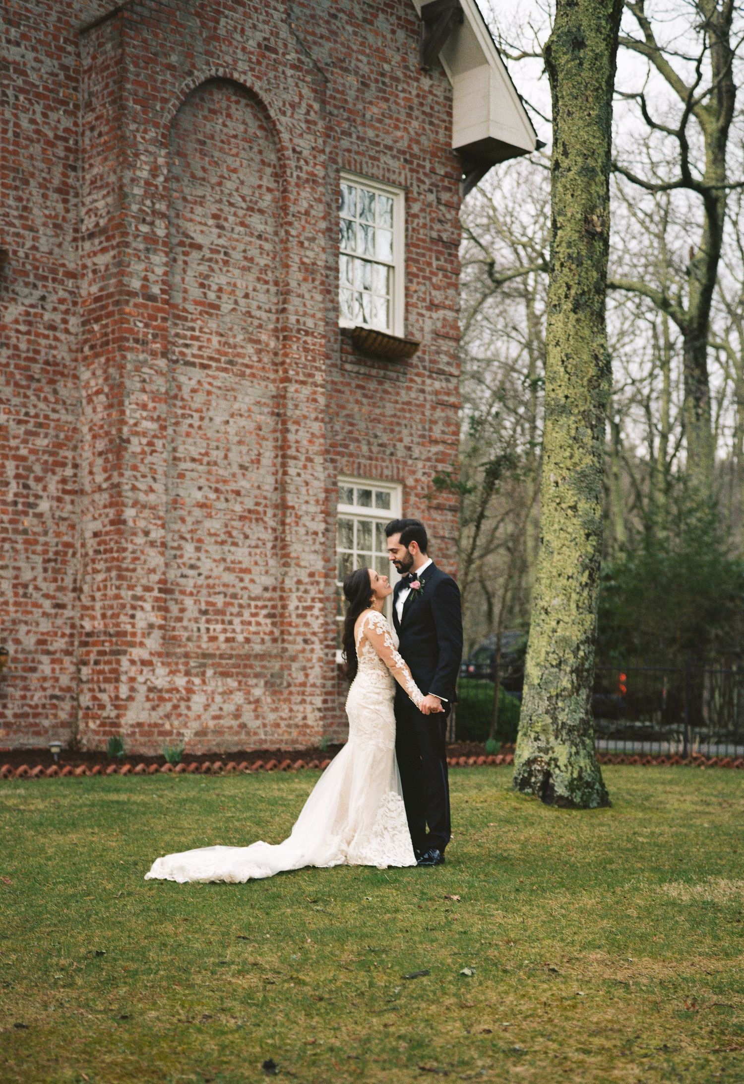Bride and groom share a romantic moment on green lawn beside historic red brick building and bare tree.