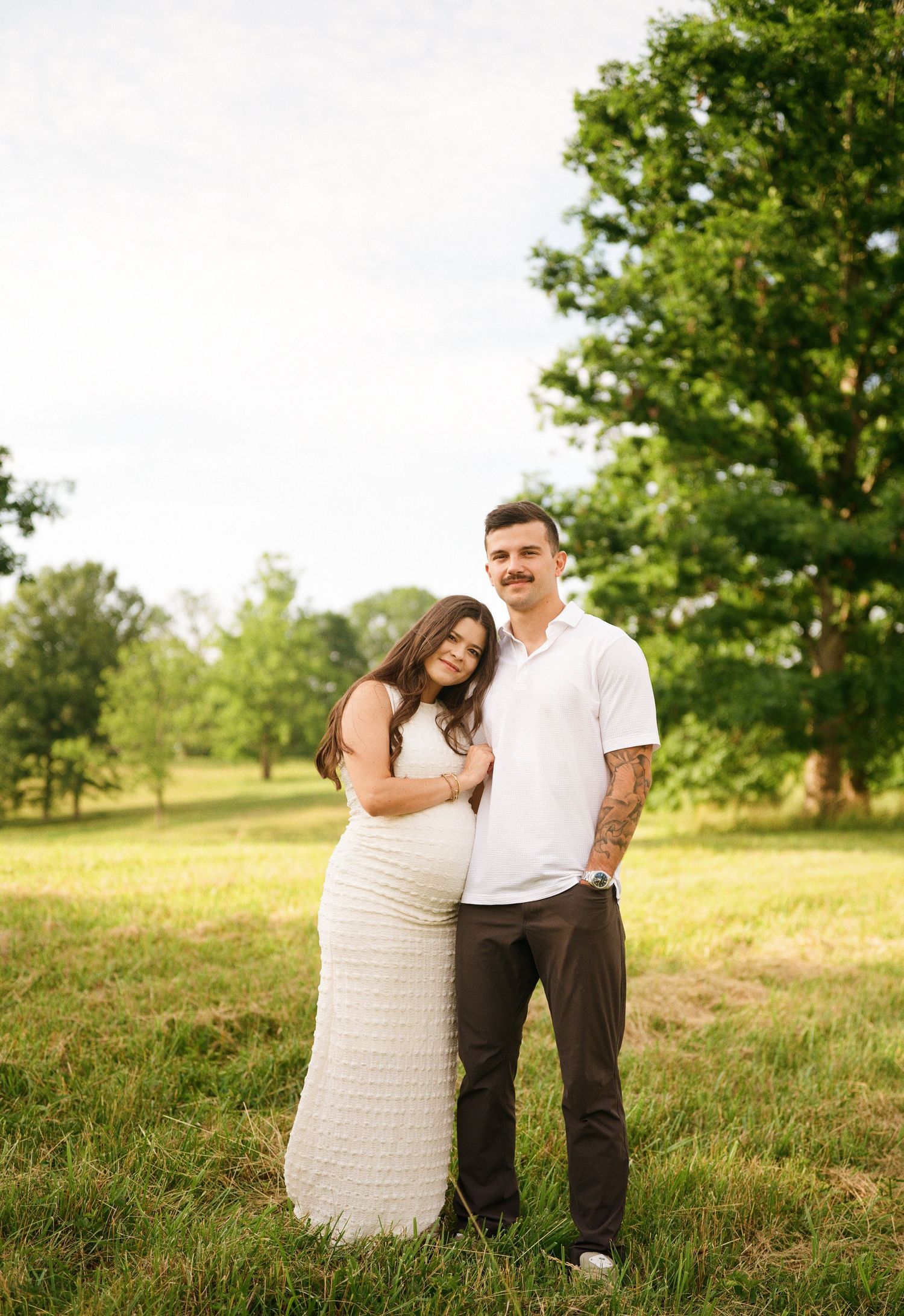 Pregnant woman in white dress leans on man in white polo shirt, posing in green field during maternity photo session.