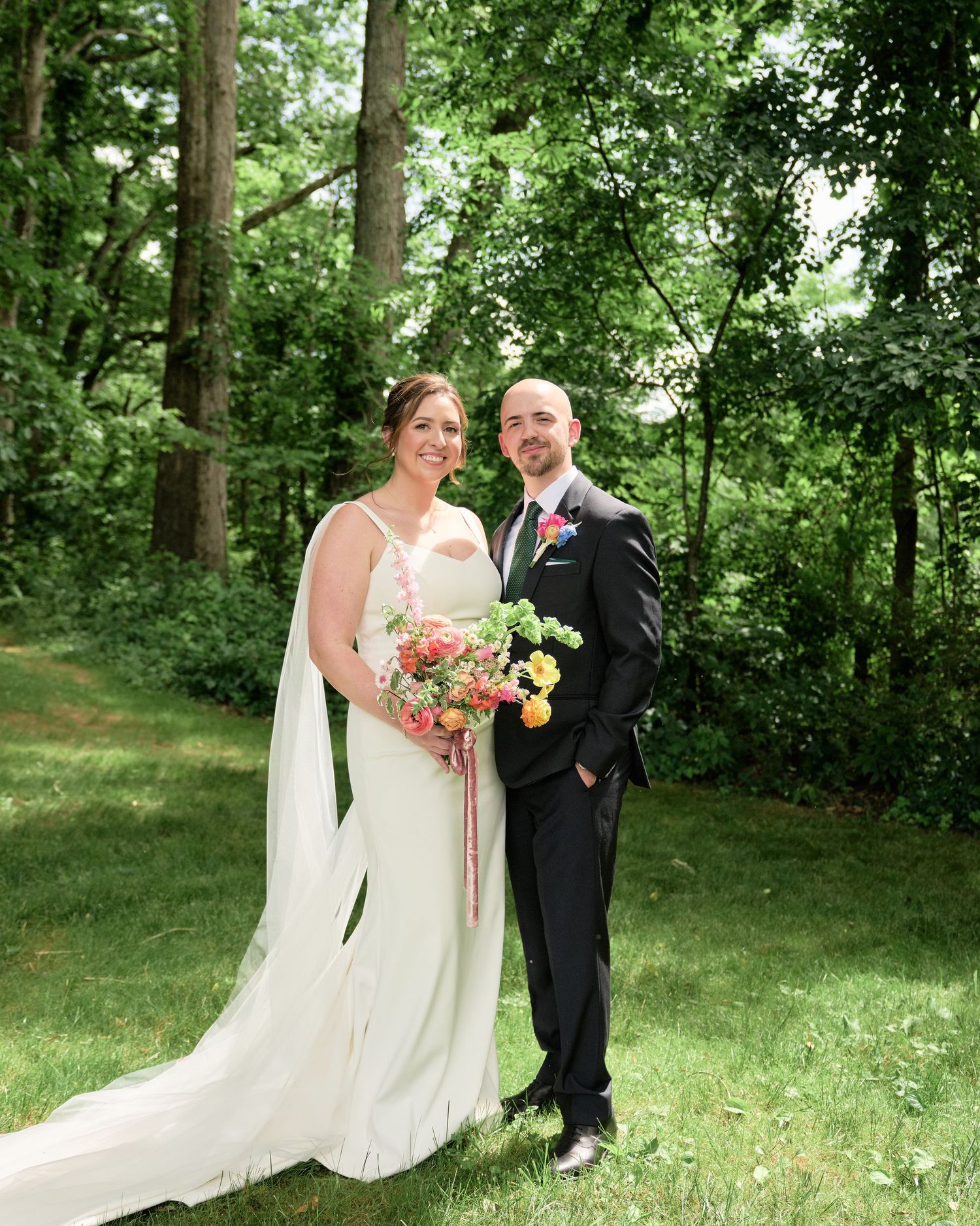 Wedding couple poses for formal portrait on grass with forest backdrop and colorful bouquet.