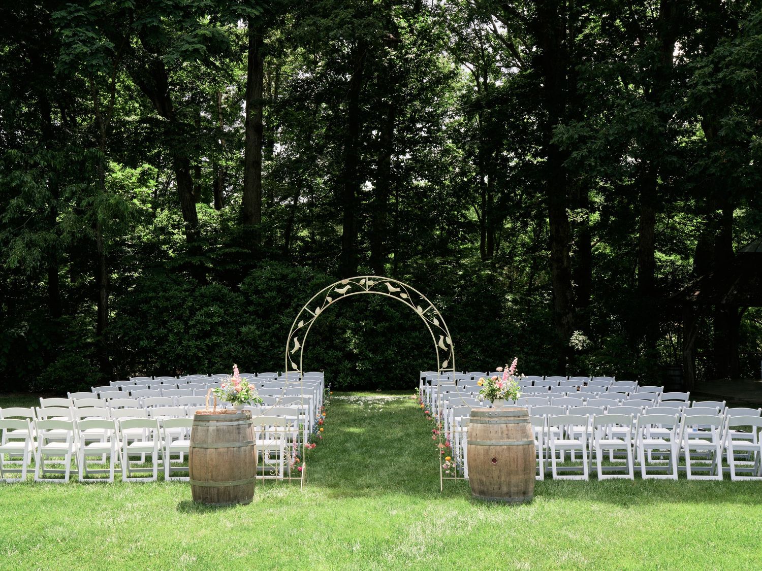 An outdoor wedding ceremony setup with white chairs and wine barrel accents under a floral arch in a forest setting.